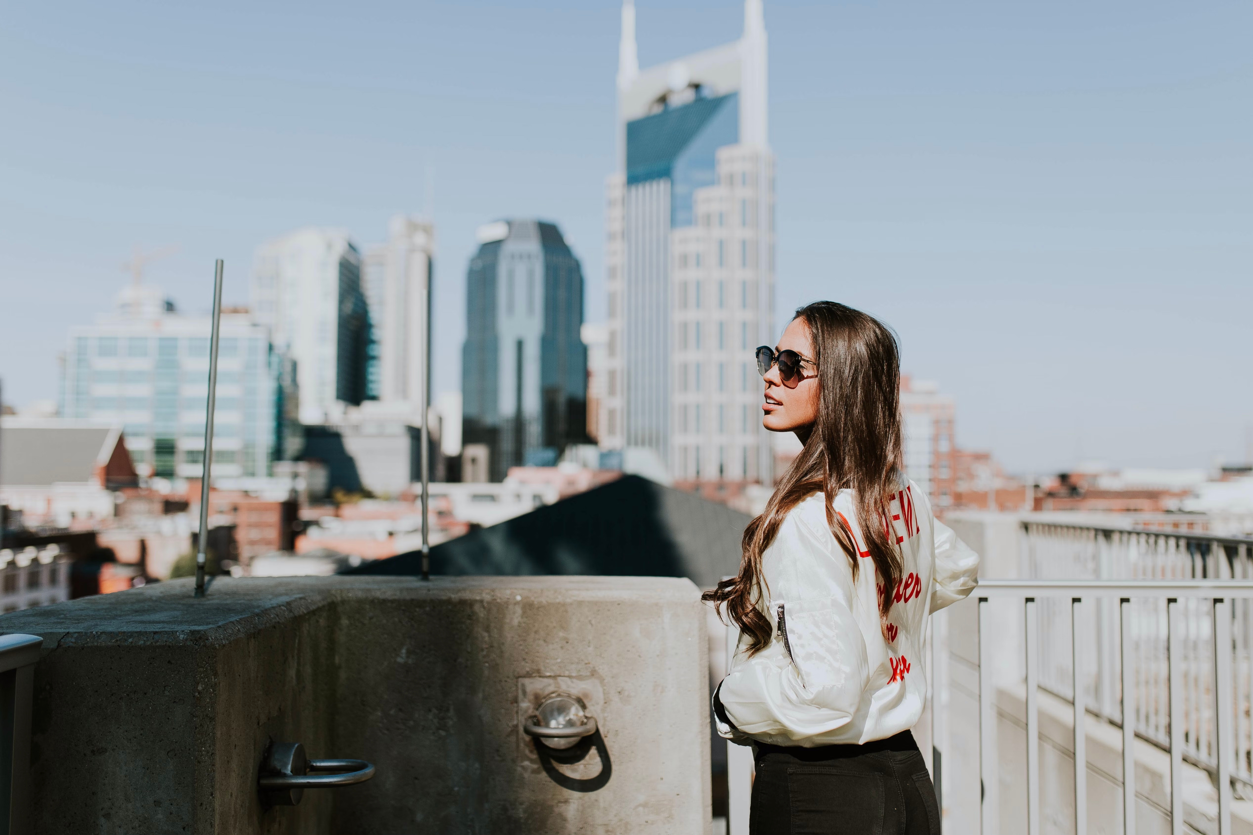 Woman with long hair and sunglasses stands on a rooftop overlooking the vibrant Nashville city skyline, featuring modern tall buildings and a clear sky.