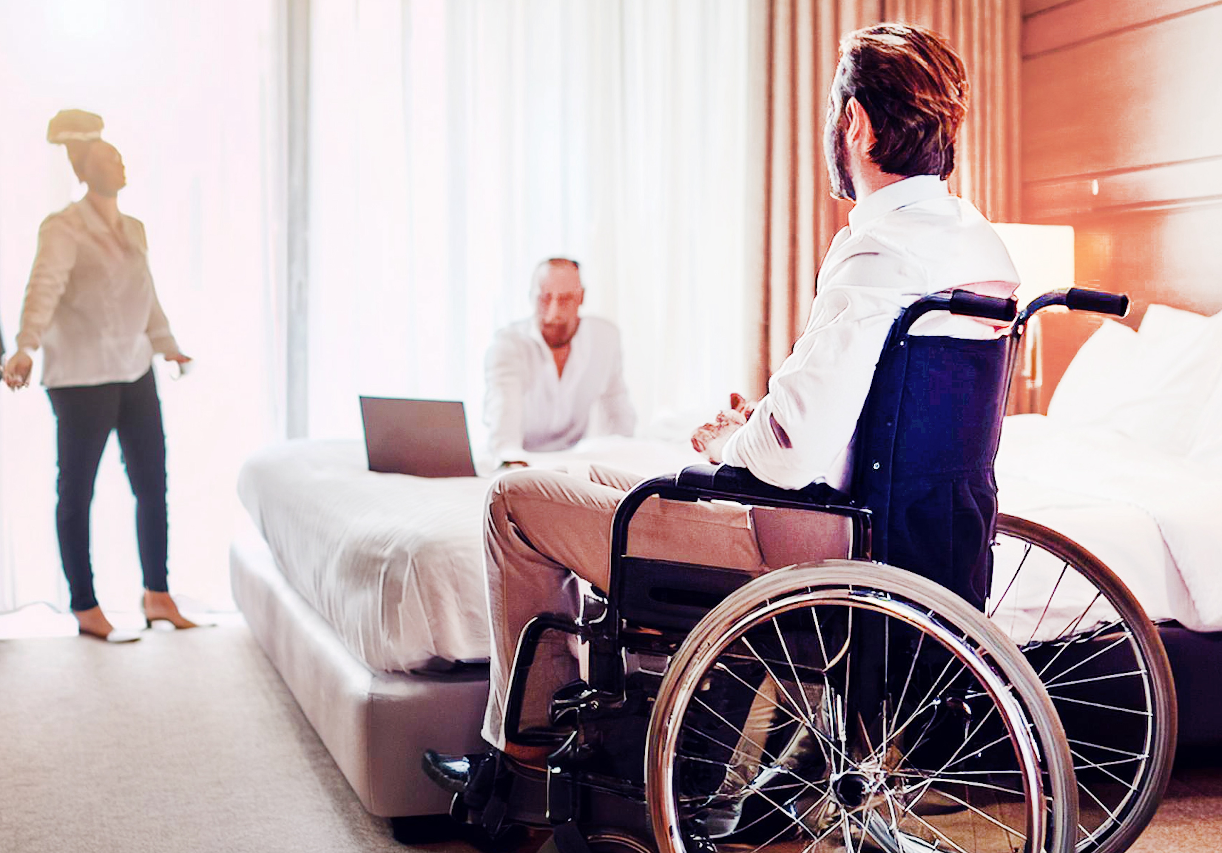 In a Nashville hotel room, a man in a wheelchair and two others engage in a lively discussion, with a laptop open on the bed.