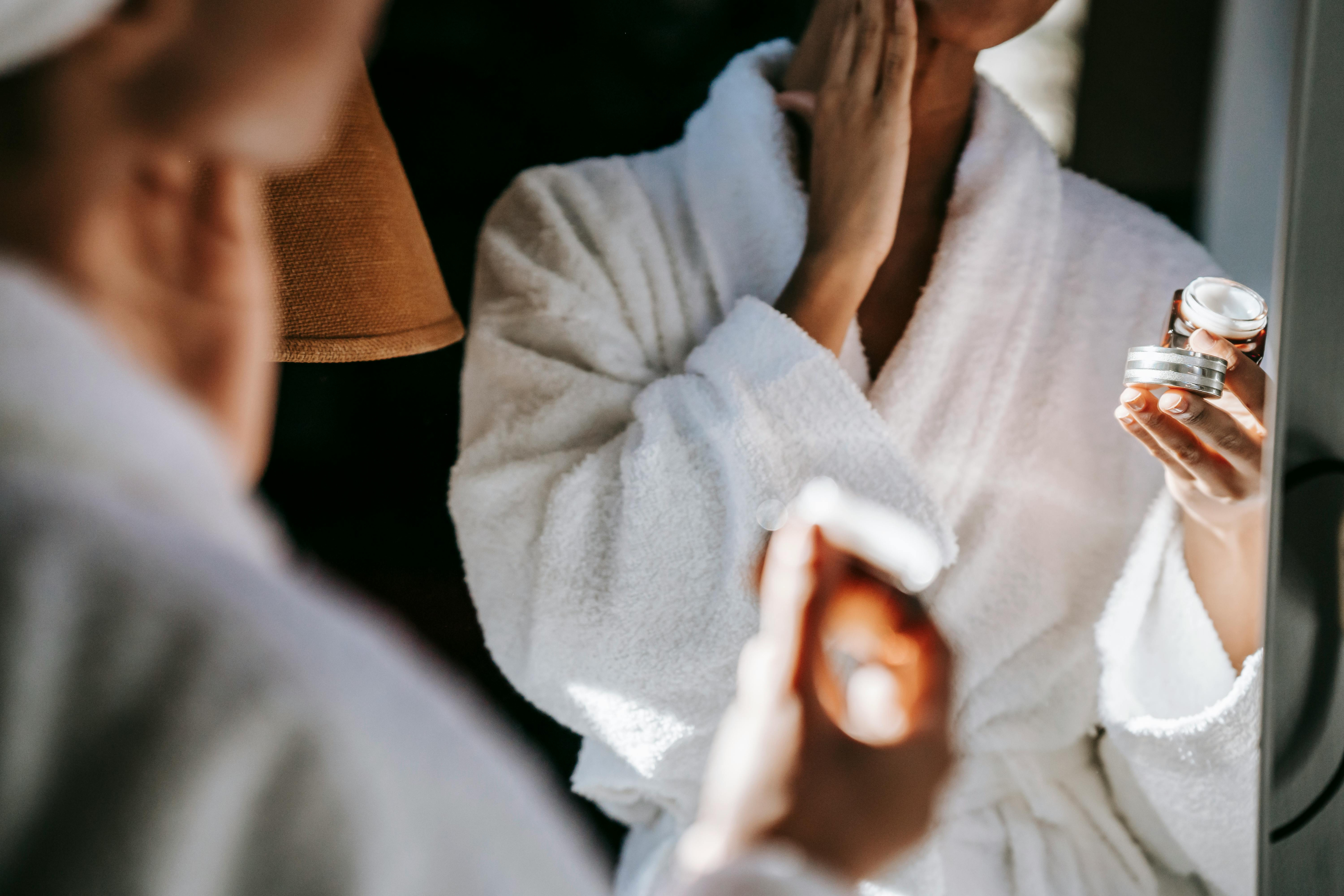 In a cozy Nashville apartment, a person in a white bathrobe applies face cream, holding a small jar, while standing in front of the mirror.