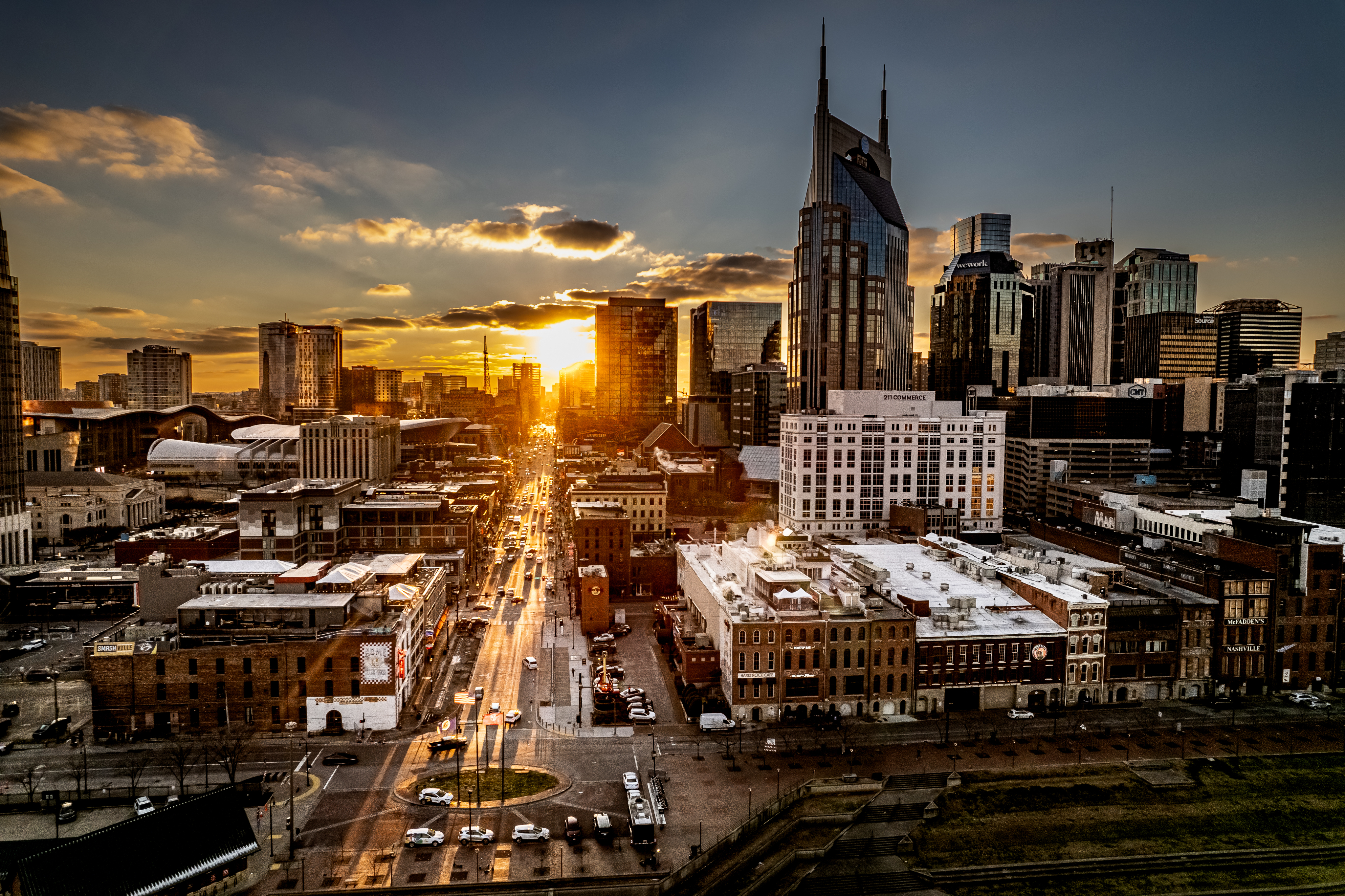 A cityscape at sunset in Nashville features a tall, pointed skyscraper in the background. The sun sets between buildings, casting a warm glow over the streets and iconic structures of Music City.