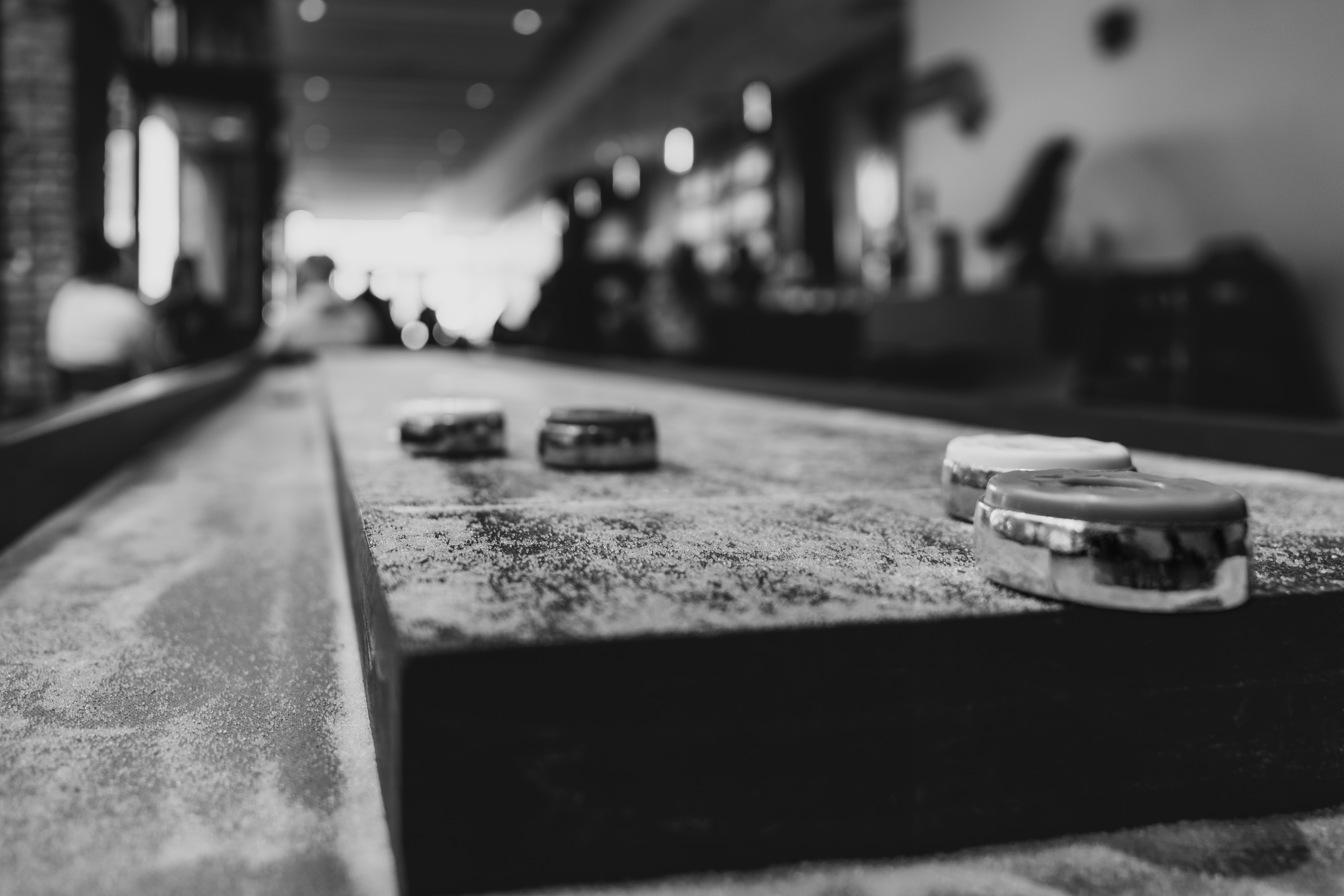 A black and white photo captures a Nashville shuffleboard table with four pucks gracefully positioned across its sandy surface, set against a blurred interior backdrop.