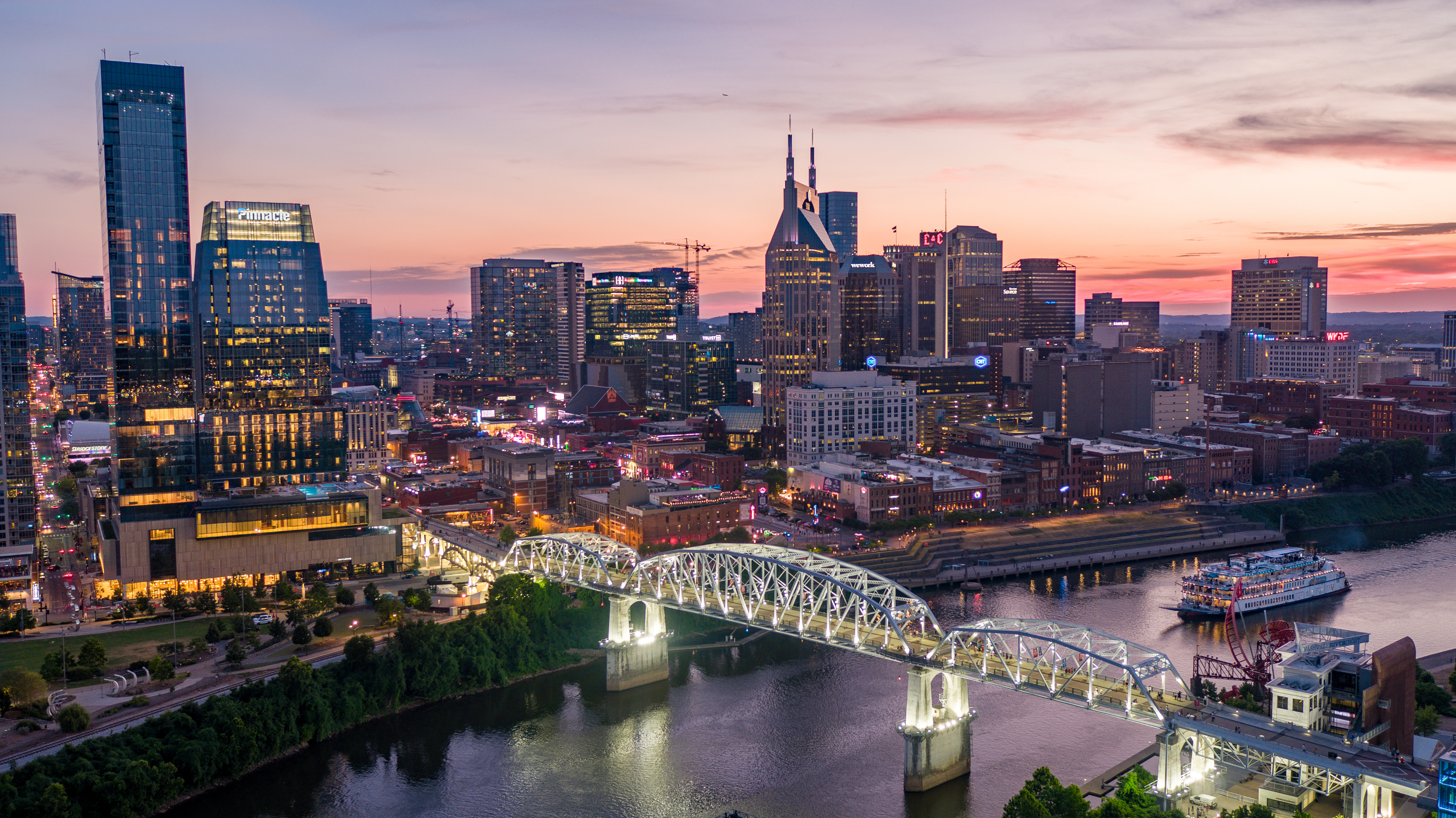 An aerial view of Nashville's skyline at sunset, showcasing a prominent bridge arching over the river, surrounded by an eclectic mix of high-rise and mid-rise buildings.