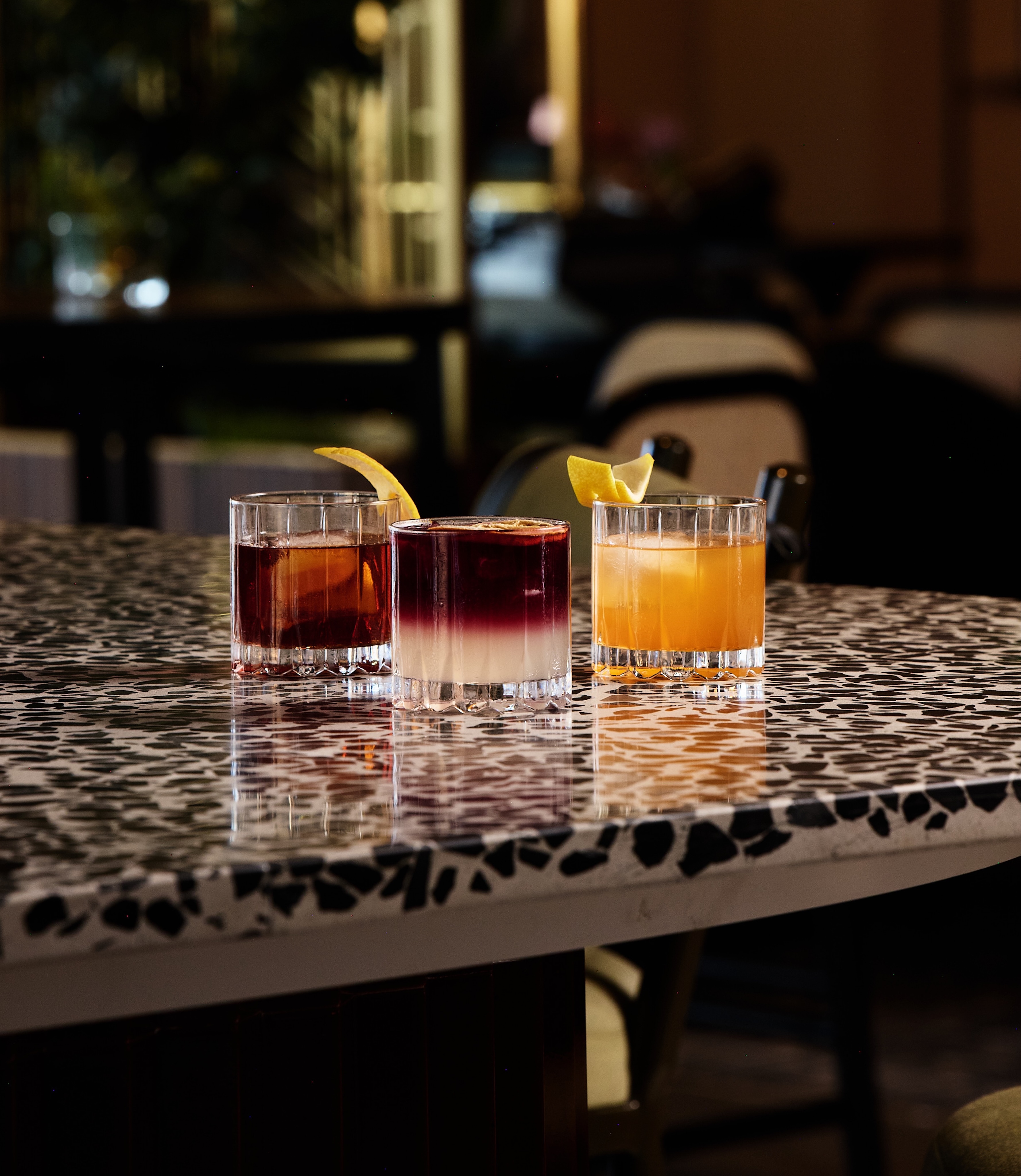 Three cocktails in short glasses, each garnished with lemon peel, are arranged on a black-and-white speckled countertop in a dimly lit bar setting—perfect for our Grand Opening. RSVP to experience the atmosphere and enjoy crafted drinks.