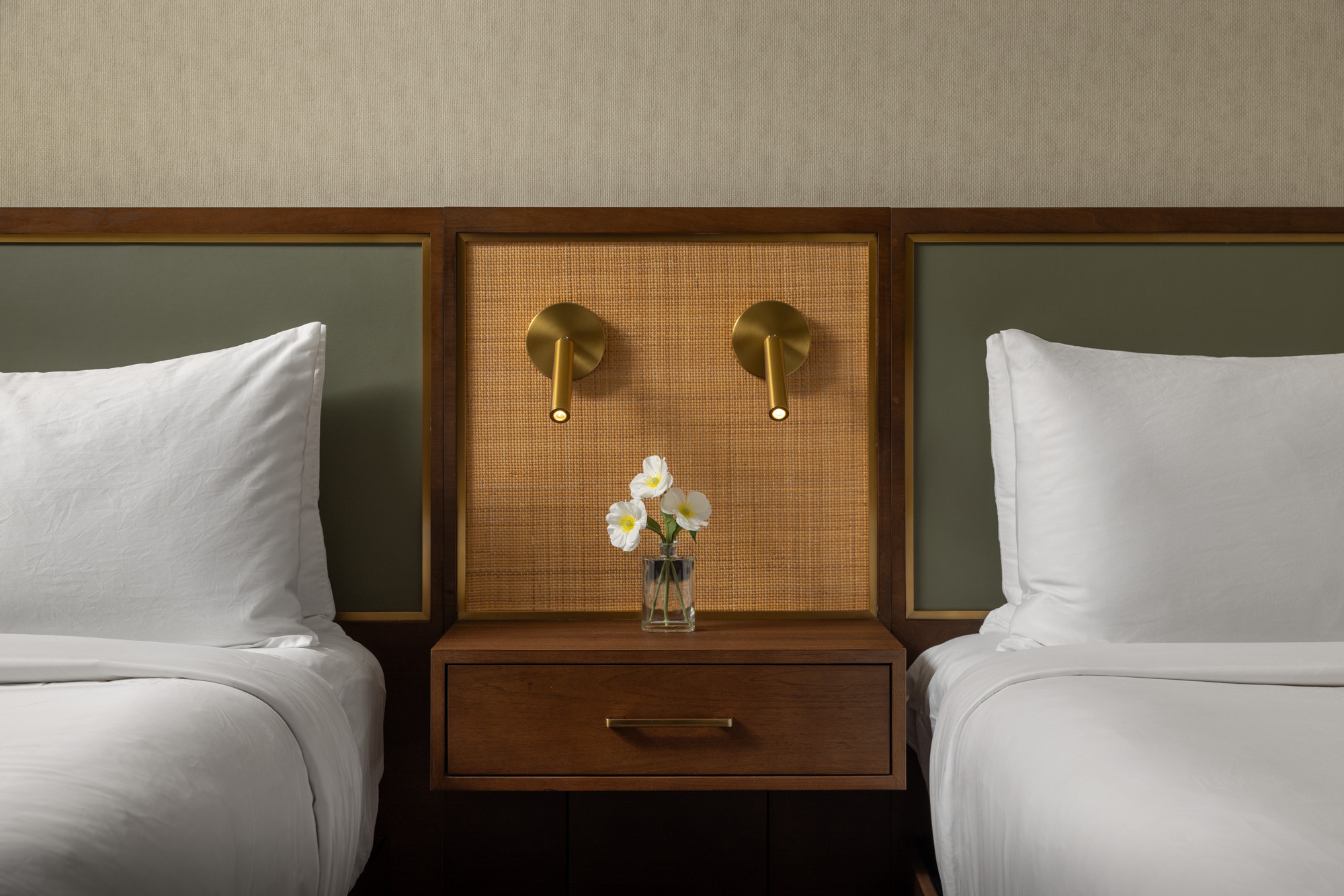 A hotel room nightstand with a small vase of white flowers, positioned between two beds with white linens and wall-mounted brass reading lights.