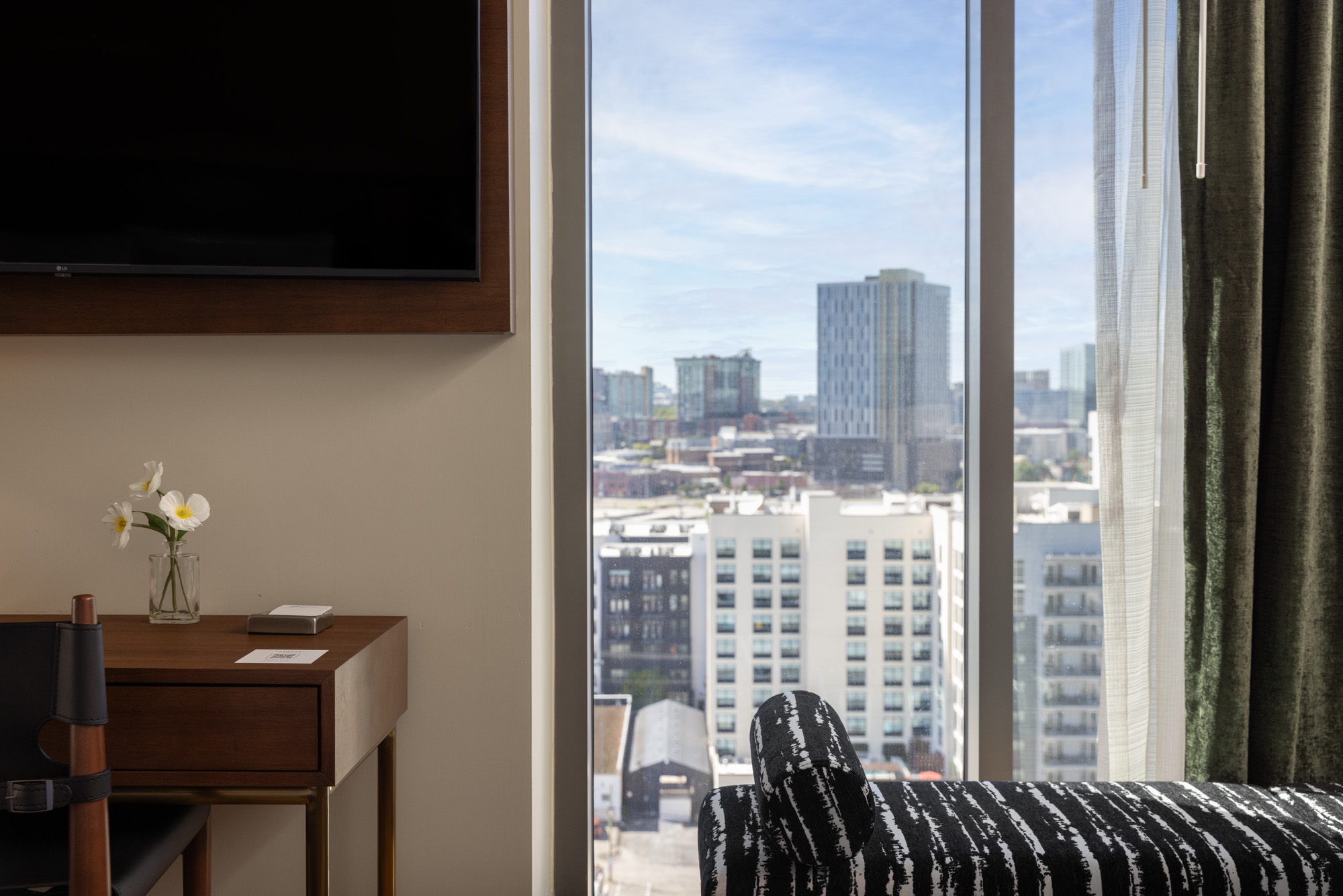 A hotel room with a desk, chair, and small vase of flowers is shown next to a large window overlooking a cityscape with tall buildings.