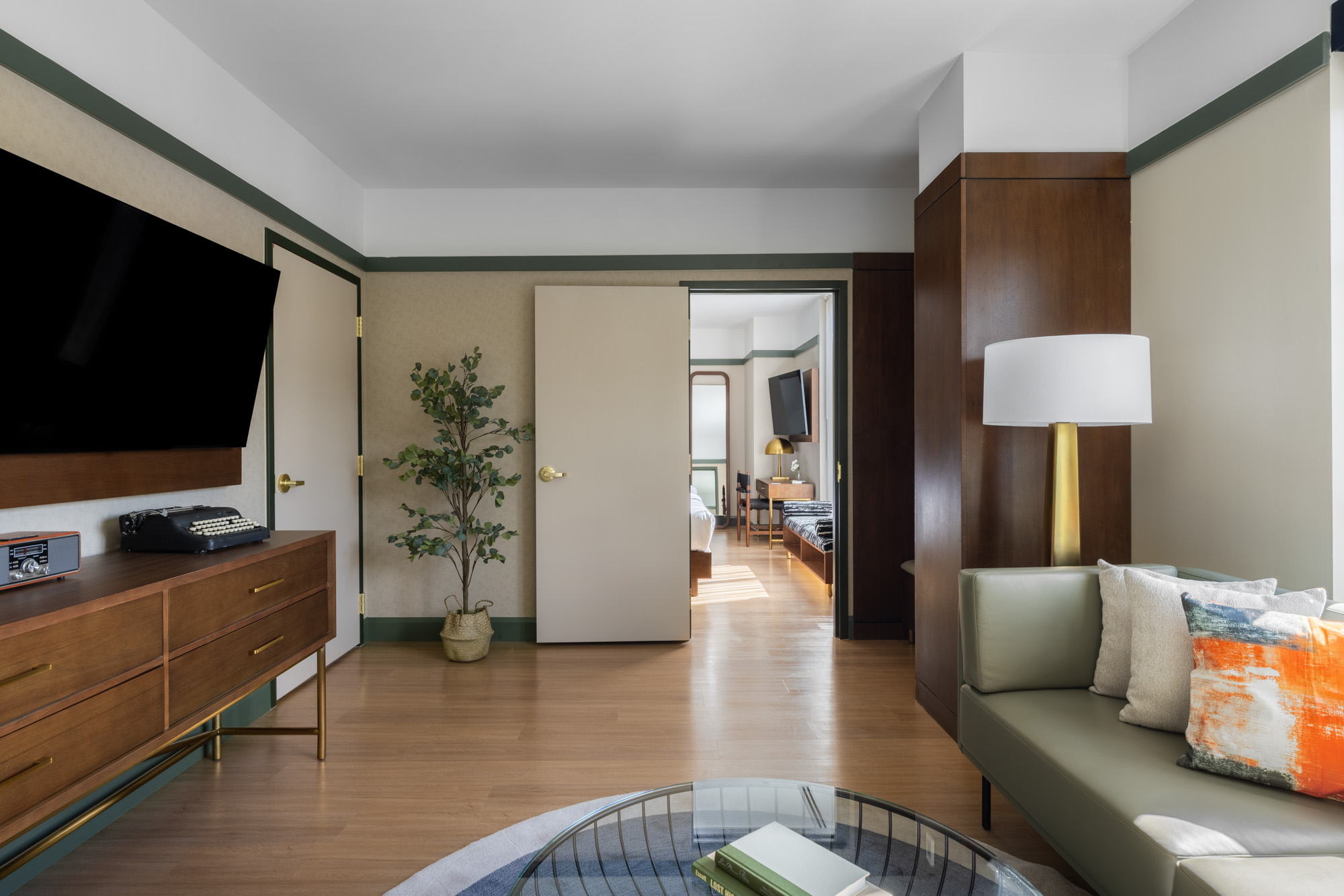 Modern living room with a TV, wooden cabinet, potted plant, round glass coffee table, green sofa, floor lamp, and an open door leading to a bedroom.