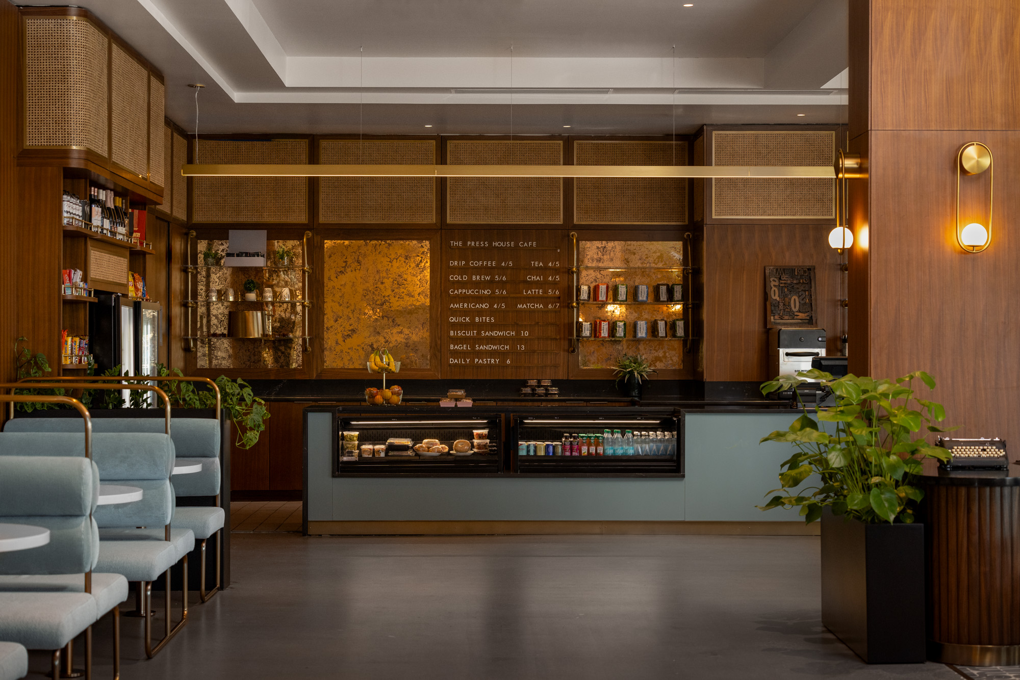 Modern café interior with wooden accents, blue seating, menu board on the wall, pastry display, shelves with products, and potted plants.