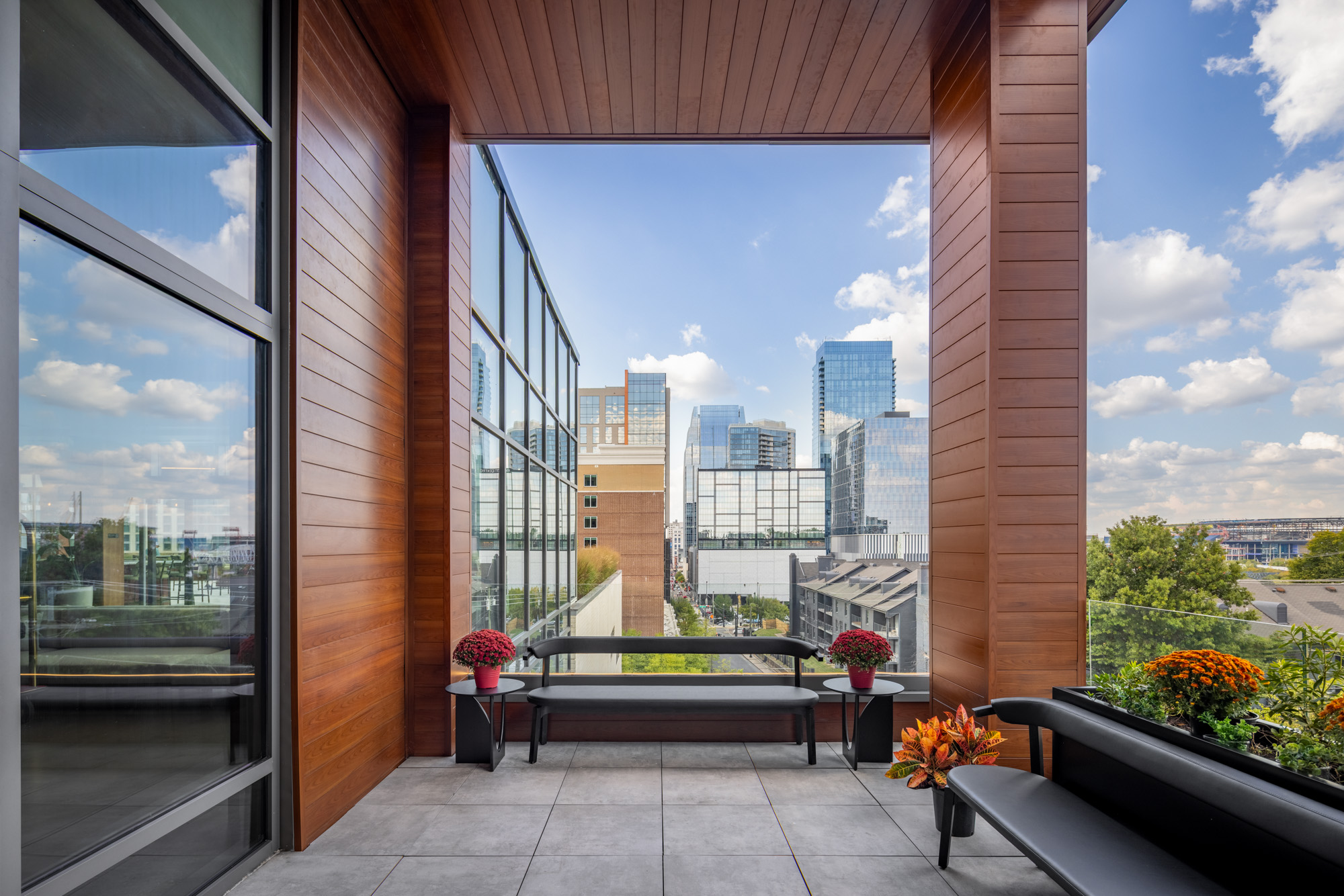 Modern outdoor balcony with wood paneling, glass walls, black benches, potted plants, and a city skyline view under a partly cloudy sky.