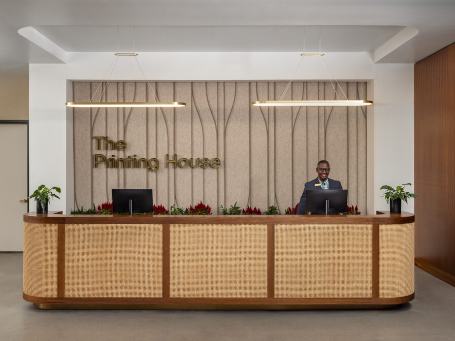 A receptionist stands behind a wooden front desk with two monitors at The Pinning House. Plants are placed on the desk and behind it.