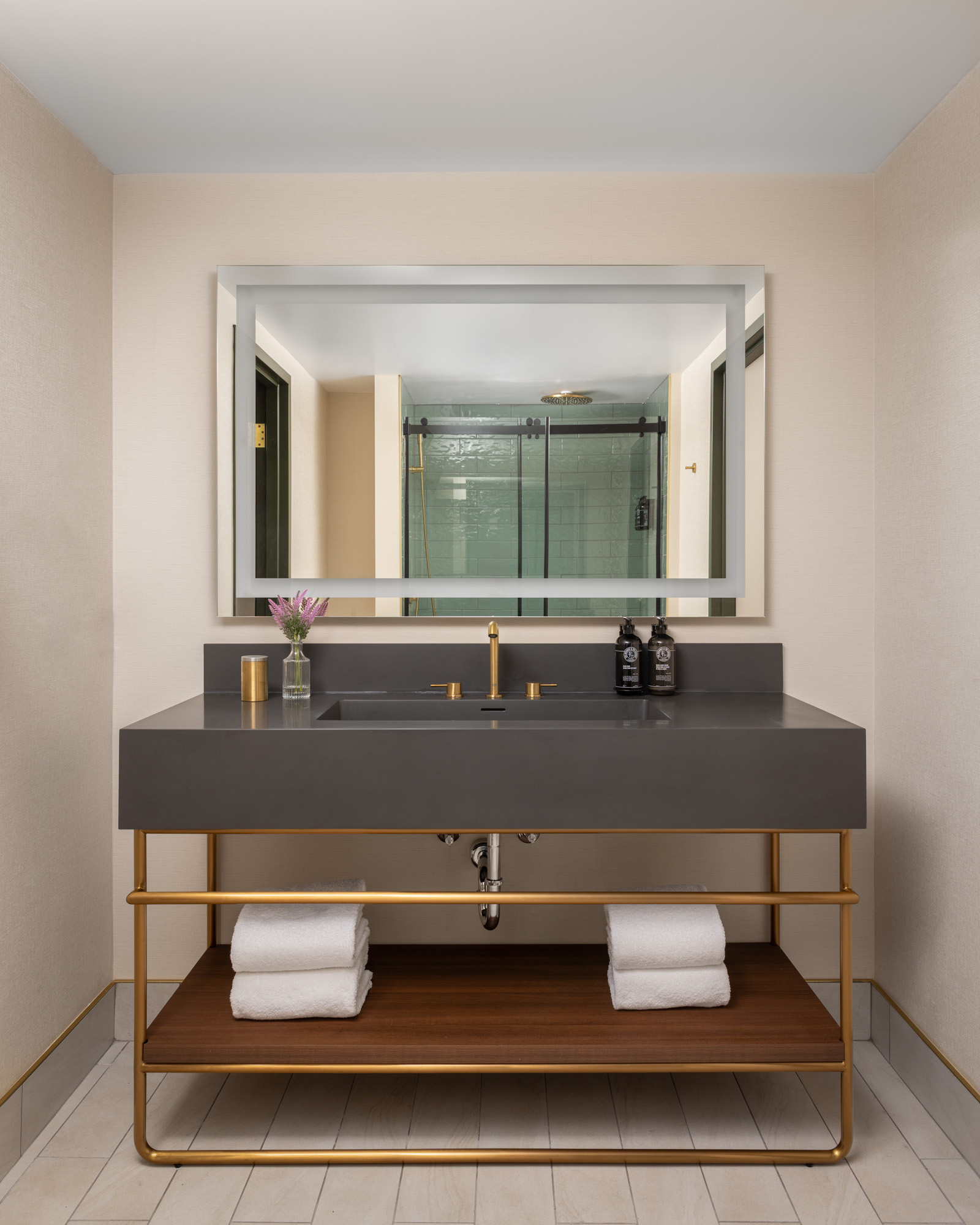 Modern bathroom with a large mirror, gray vanity with gold fixtures, two towels on a wooden shelf, and toiletries on the counter; glass shower visible in the reflection.
