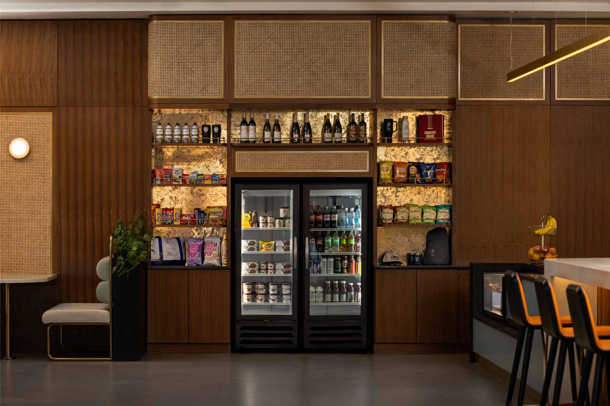 A modern self-serve snack and beverage area with two glass-door refrigerators, shelves of snacks and drinks, and wood paneling.