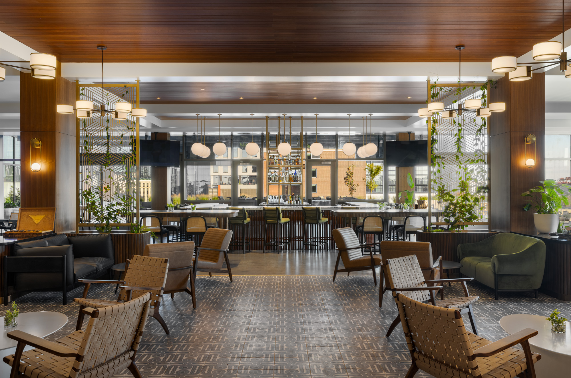 Modern hotel lobby with woven chairs, patterned floor, large windows, indoor plants, and a central bar area illuminated by pendant lights.