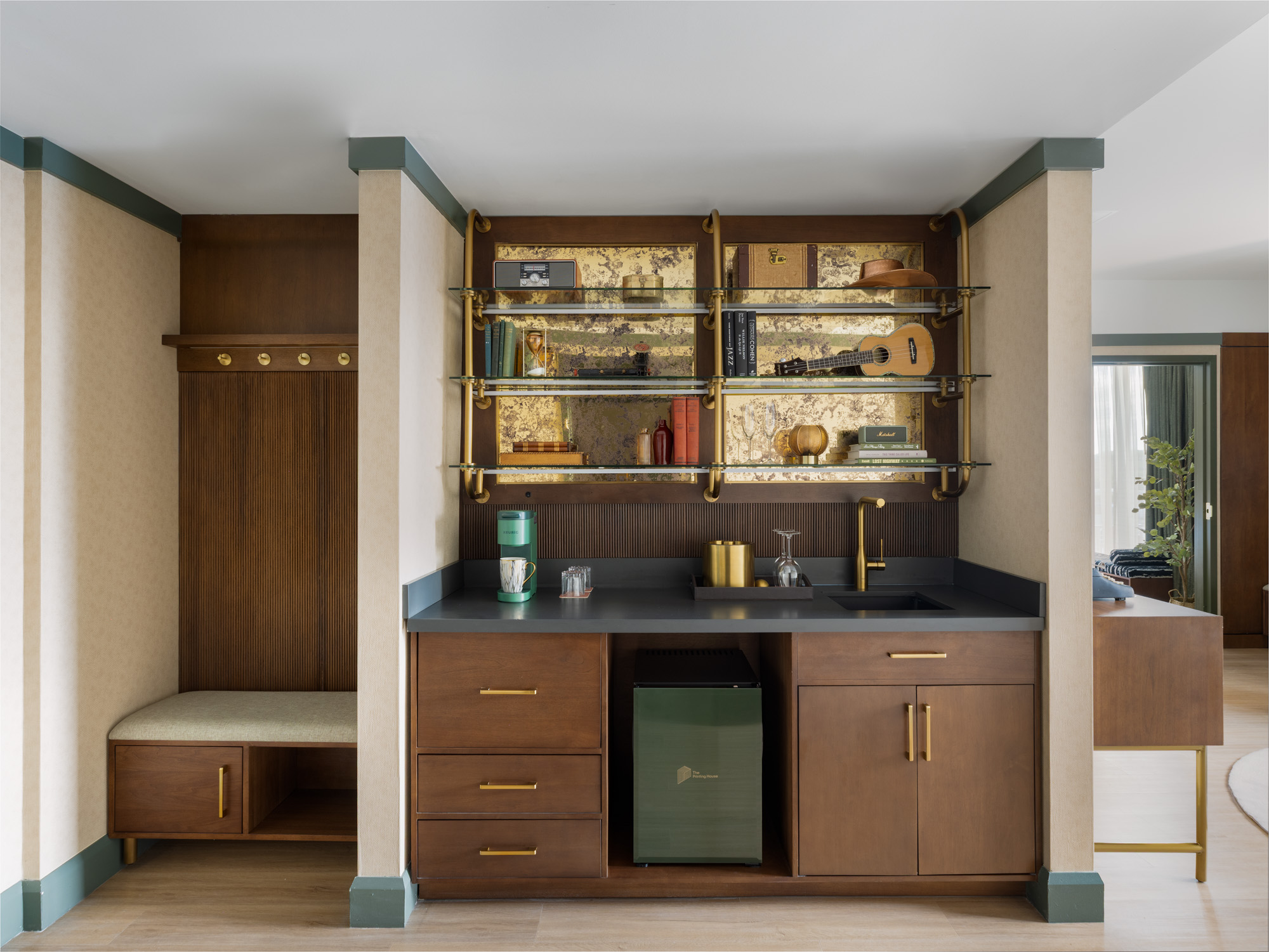 Modern kitchenette with dark wood cabinets, a small green fridge, sink, coffee maker, decorative wall shelving, and a bench with coat hooks on the left side.