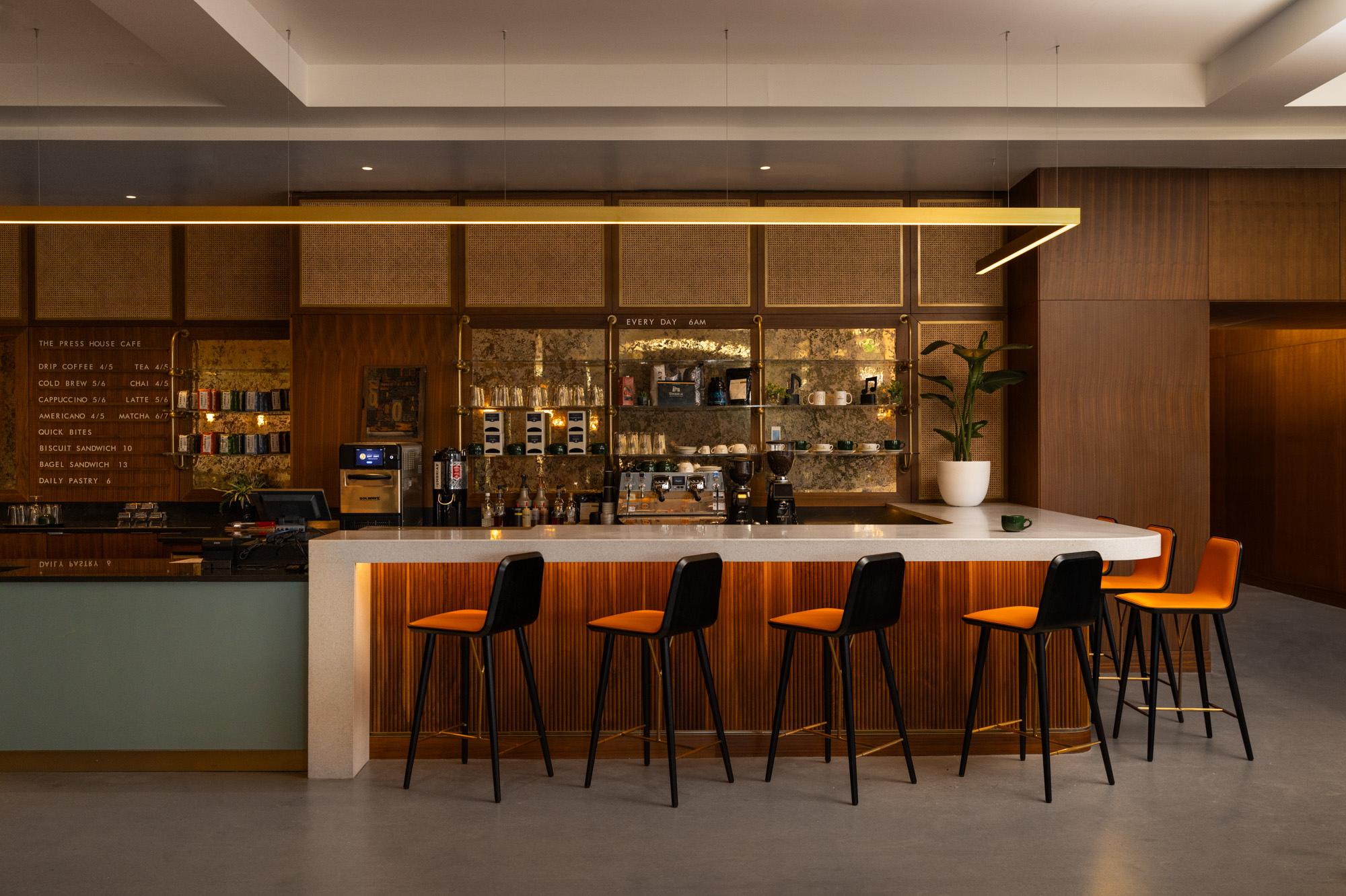 A modern café bar with a white countertop, orange and black bar stools, a coffee machine, menu boards, and warm lighting. A potted plant sits at one end of the counter.