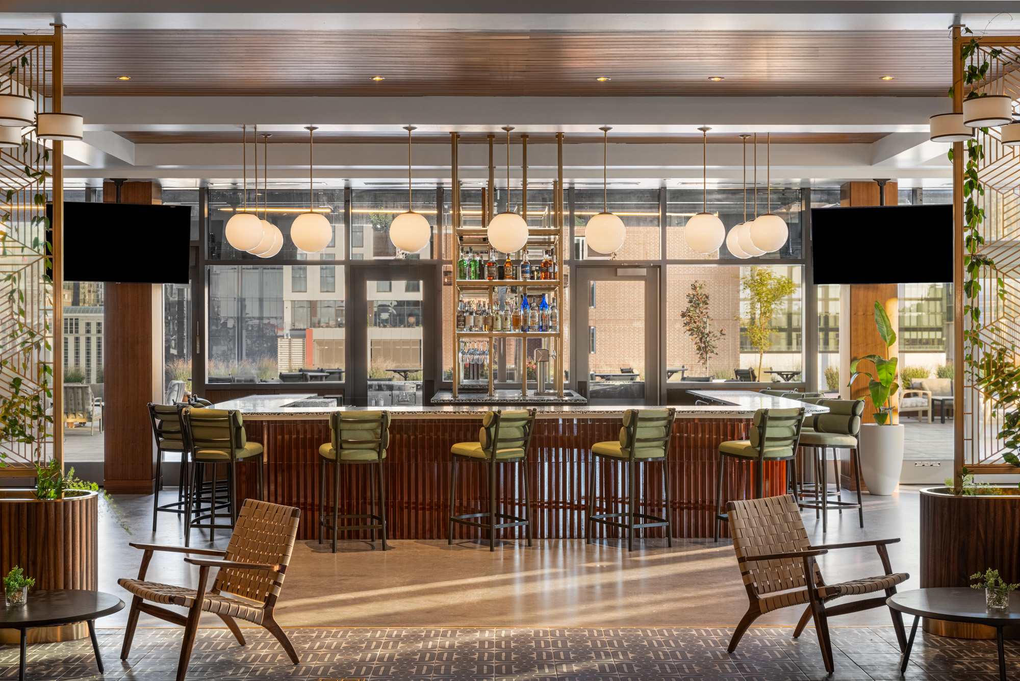 A modern hotel bar with green barstools, globe pendant lights, and a variety of bottles on shelves behind the counter, surrounded by lounge chairs and plants.