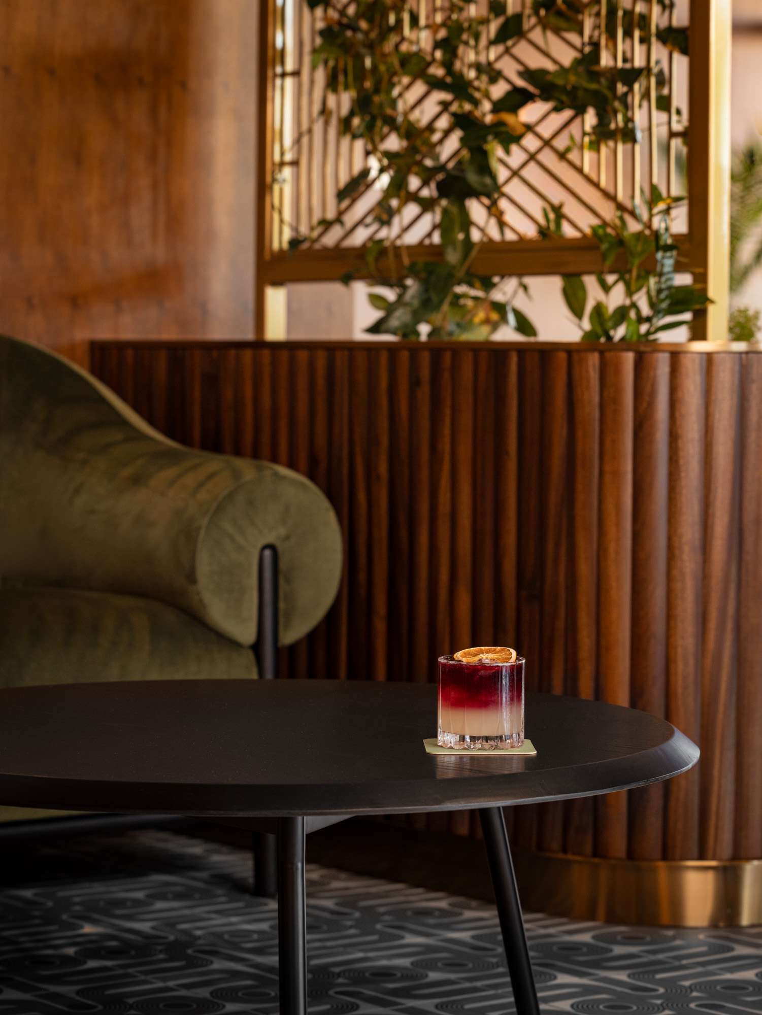 A cocktail with a citrus garnish sits on a black round table in a modern lounge with a green velvet chair and wood-paneled decor.