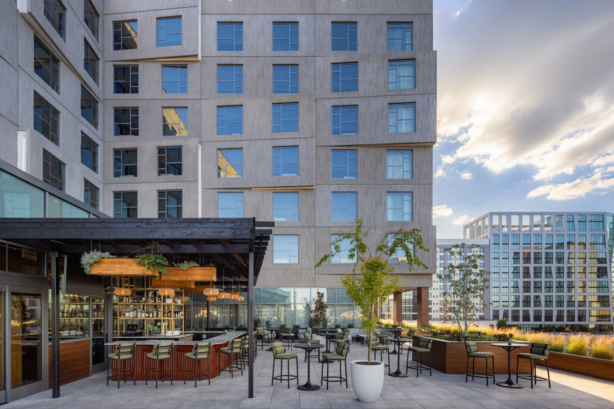 Outdoor patio with bar seating, tables, and modern decor, adjacent to a contemporary multi-story building with large windows and landscaping.