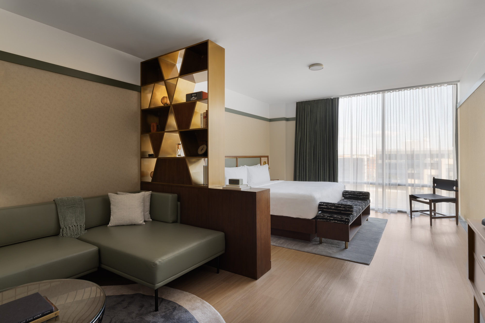 A modern hotel room with a double bed, green sectional sofa, partition shelving, a round table, large window with sheer curtains, and a chair by the window.
