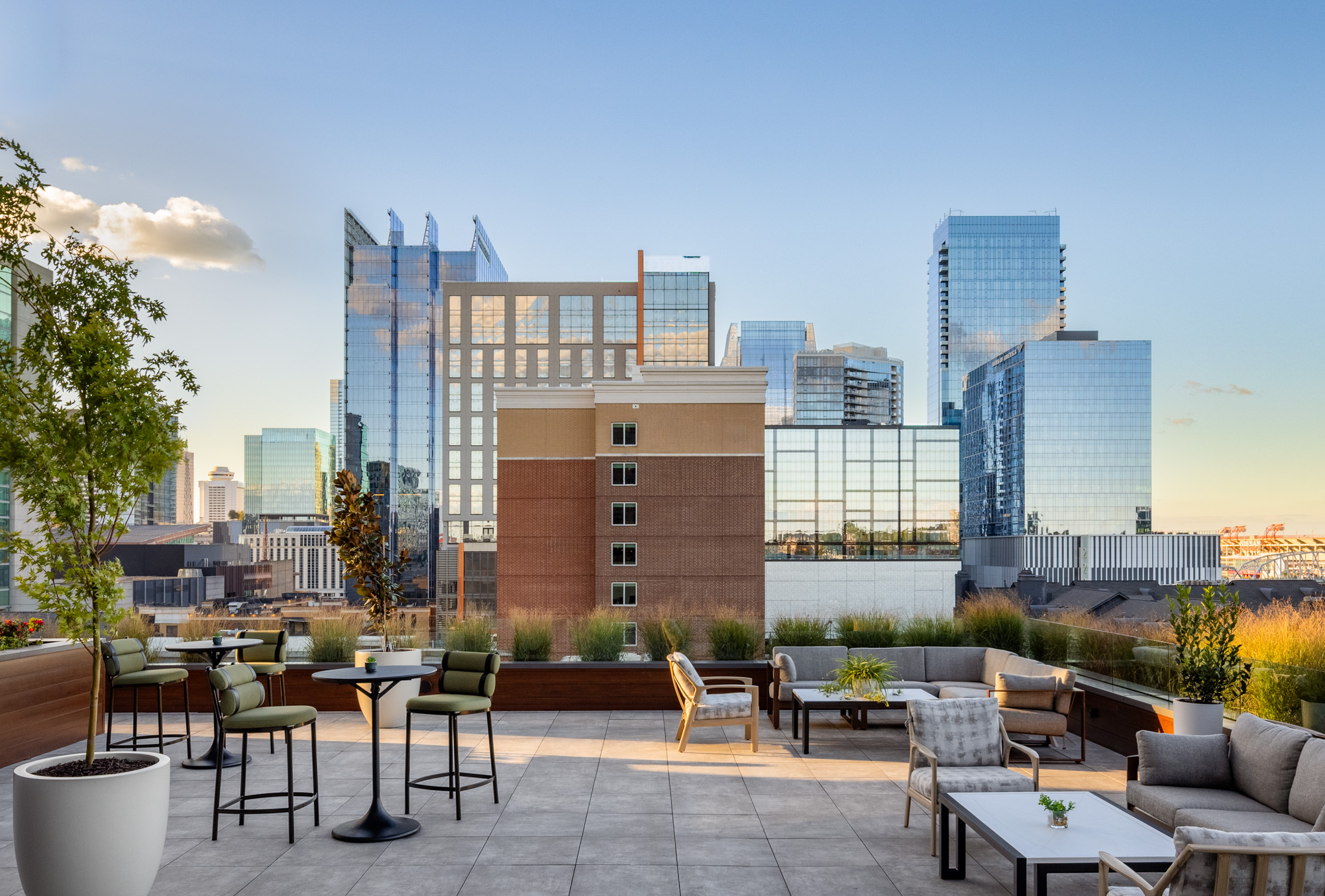 Outdoor rooftop patio with modern seating, tables, and potted plants, overlooking a cityscape of tall glass buildings under a clear sky.