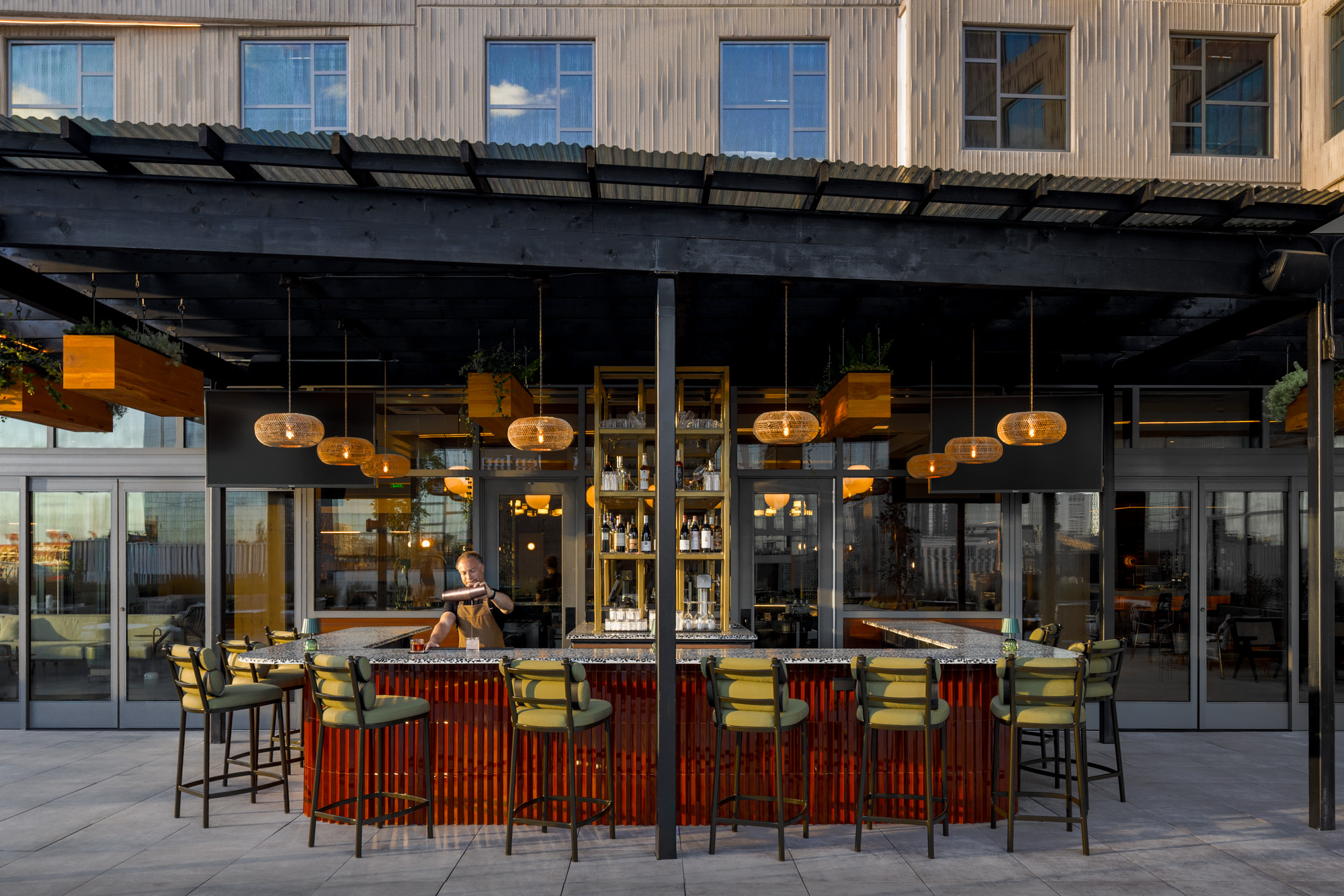 Outdoor bar with modern decor and overhead lighting, empty except for one person sitting at the counter reading, with shelves of bottles behind the bar and a building in the background.