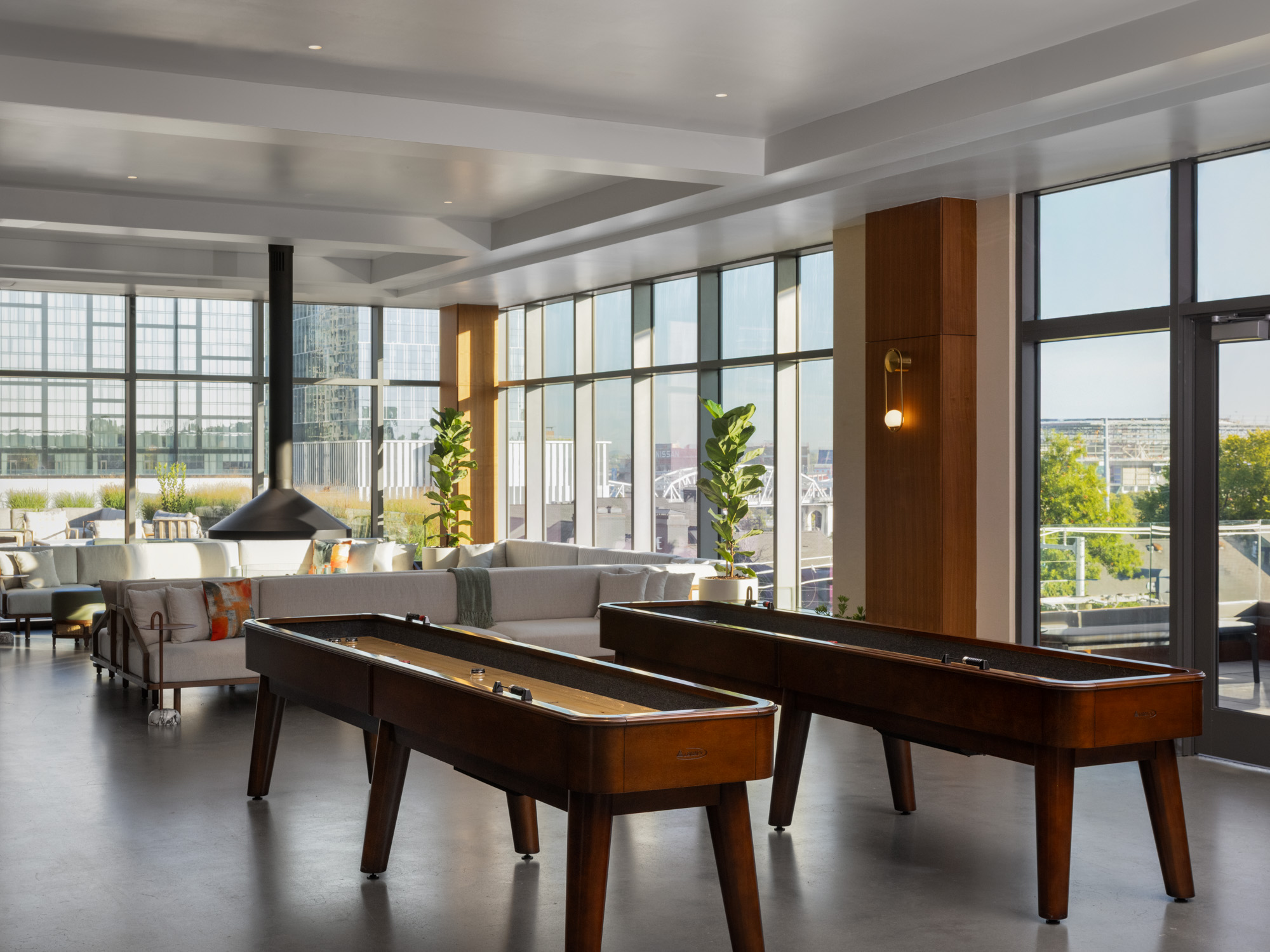 A modern lounge with two shuffleboard tables, large windows, indoor plants, sofas, and access to an outdoor balcony with city views.