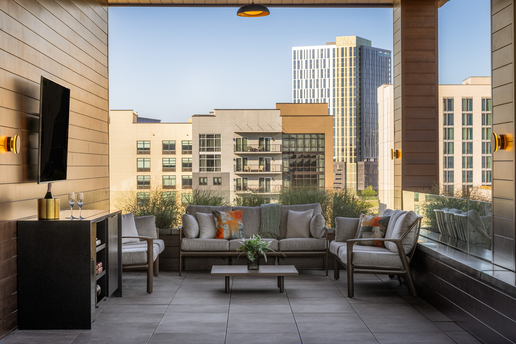 Modern outdoor patio with cushioned seating, coffee table, and wall-mounted TV, overlooking city buildings on a clear day.