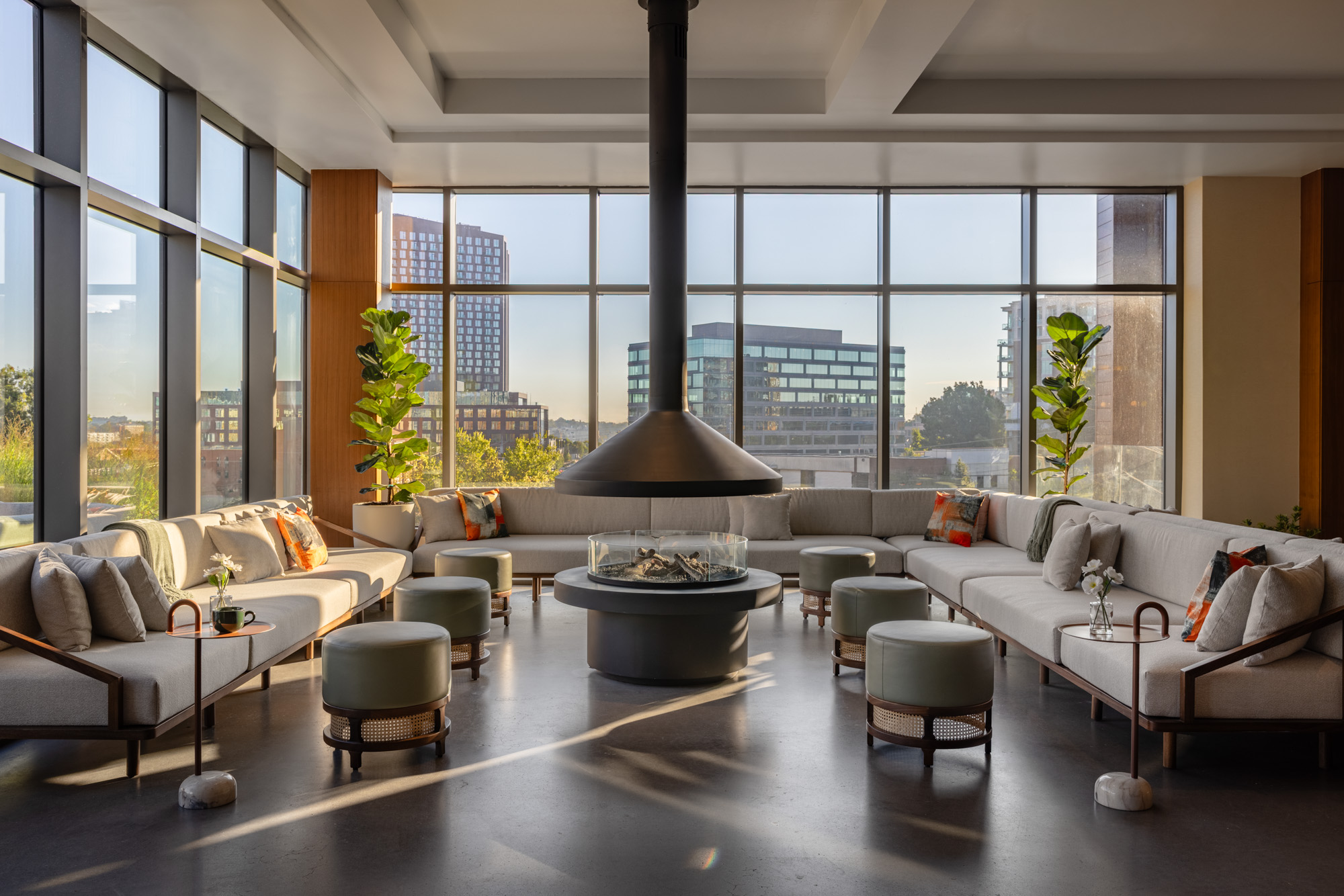 Modern lounge with large windows, neutral sofas arranged in a U-shape, green ottomans, indoor plants, and a central suspended fireplace. City buildings visible outside.