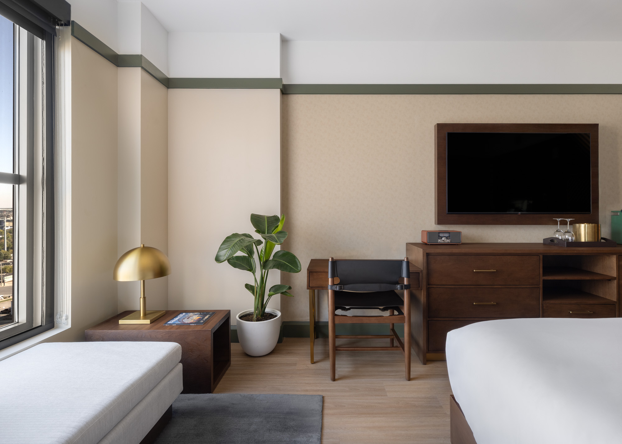 A hotel room corner with a bed, wooden dresser, mounted TV, black chair, potted plant, gold table lamp, and large window with daylight coming in.