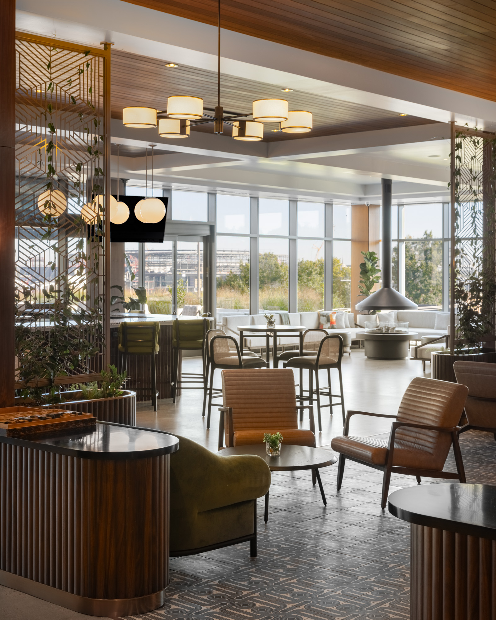 Modern lounge area with wooden furniture, geometric partitions, overhead lighting, large windows, and a central fireplace; outdoor greenery visible through the windows.