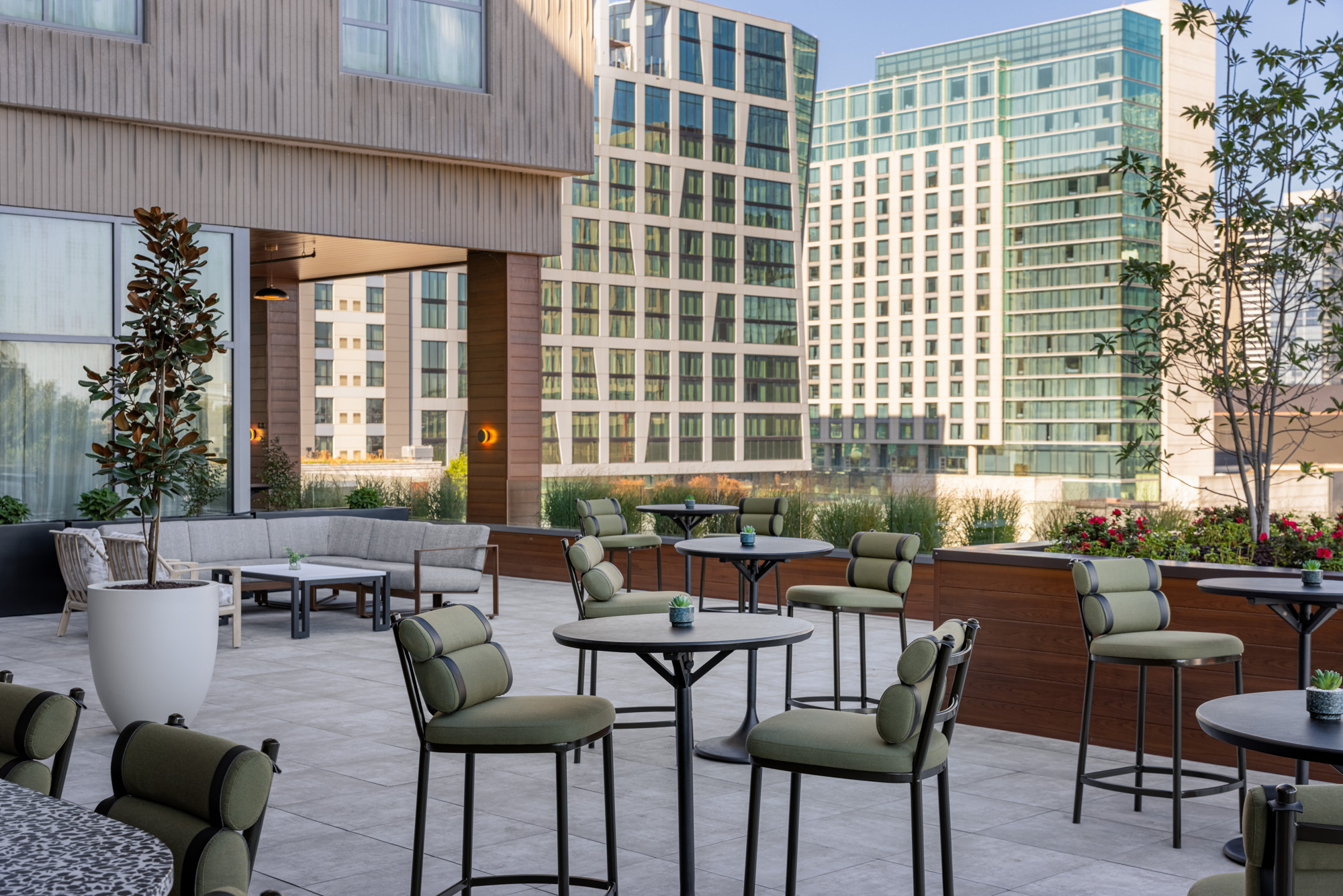 Modern outdoor patio with green cushioned chairs and round tables, surrounded by potted plants and tall glass office buildings in the background.