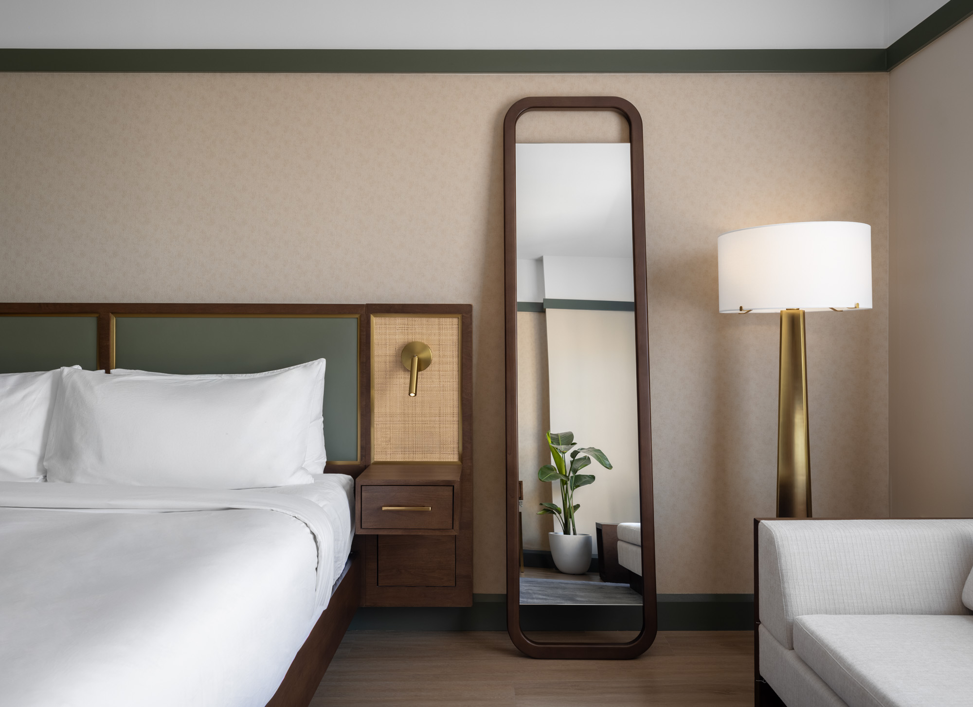 A hotel room with a neatly made bed, wall-mounted reading light, tall mirror, floor lamp, potted plant, and a white sofa.