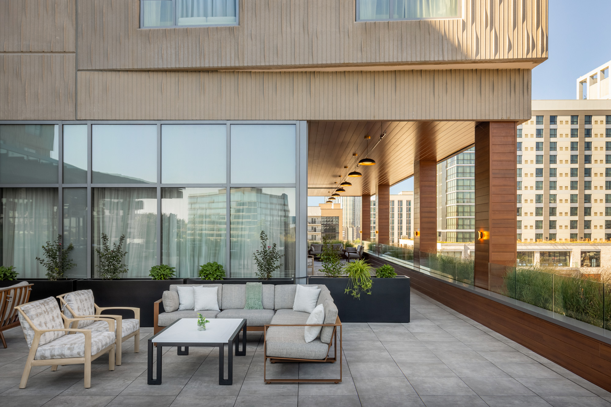 Outdoor patio with modern seating area, a sofa, chairs, coffee table, potted plants, and overhead lights, surrounded by tall buildings and glass windows.