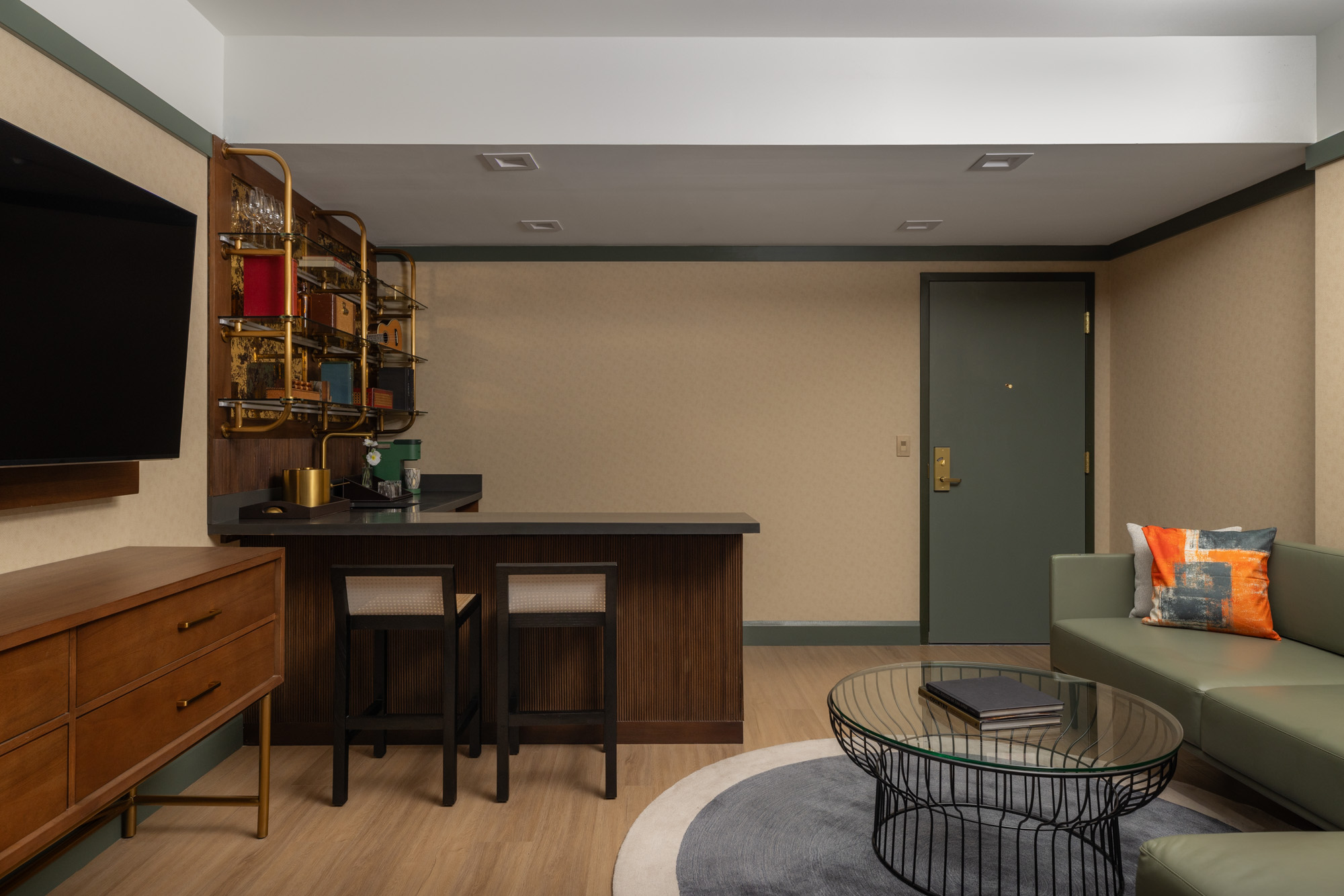 A modern hotel room bar area with two stools, a wall-mounted shelf with glassware, a green door, a TV, a green sofa, and a round coffee table on a rug.