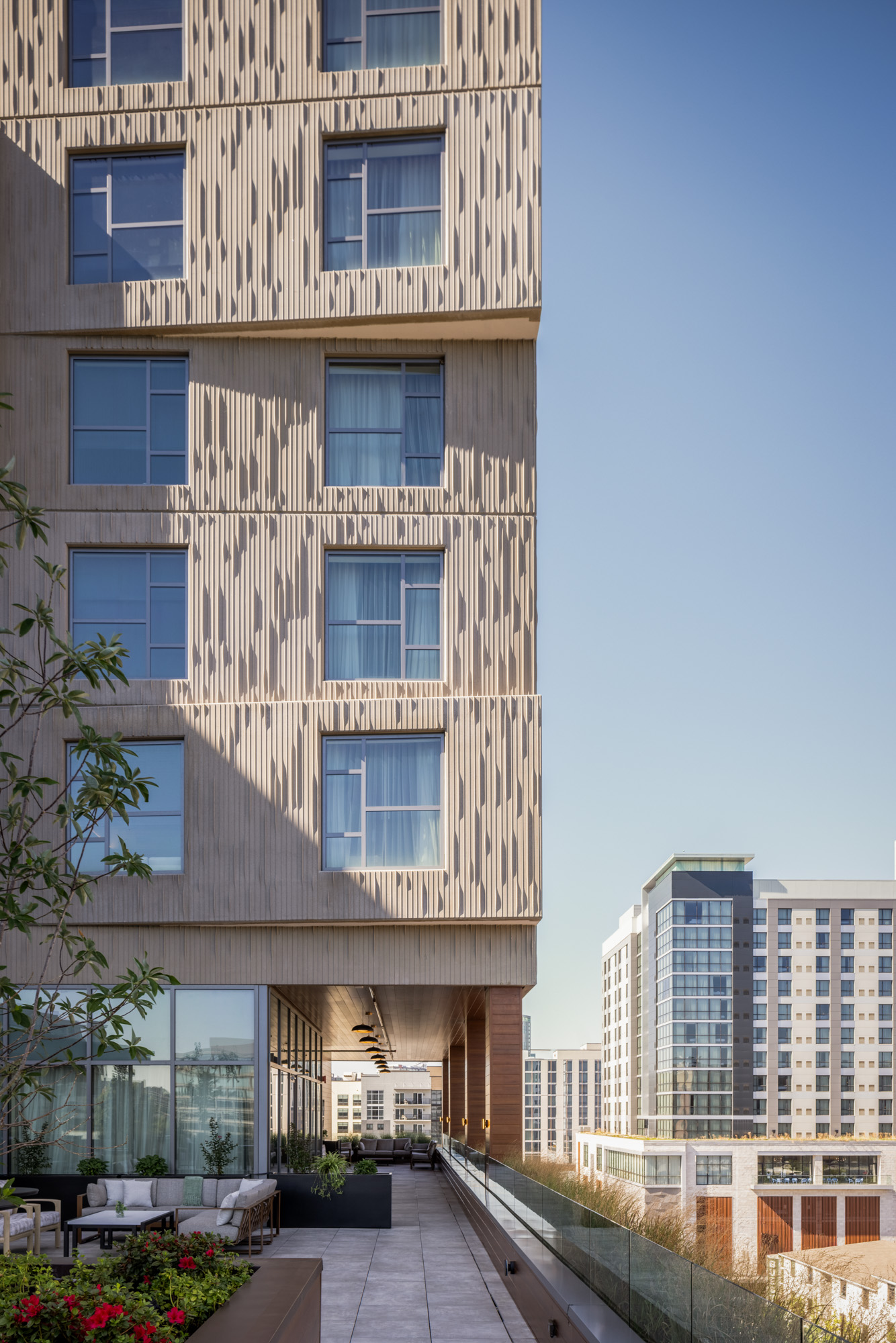 A modern multi-story building with textured facade, large windows, and a rooftop terrace overlooking other contemporary buildings under a clear sky.