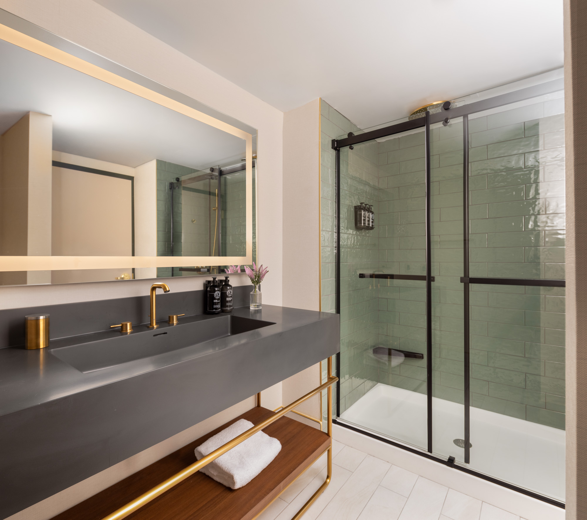Modern bathroom with a large mirror, gray sink with gold fixtures, glass-enclosed shower, light green tiles, and wooden shelf with a folded towel.
