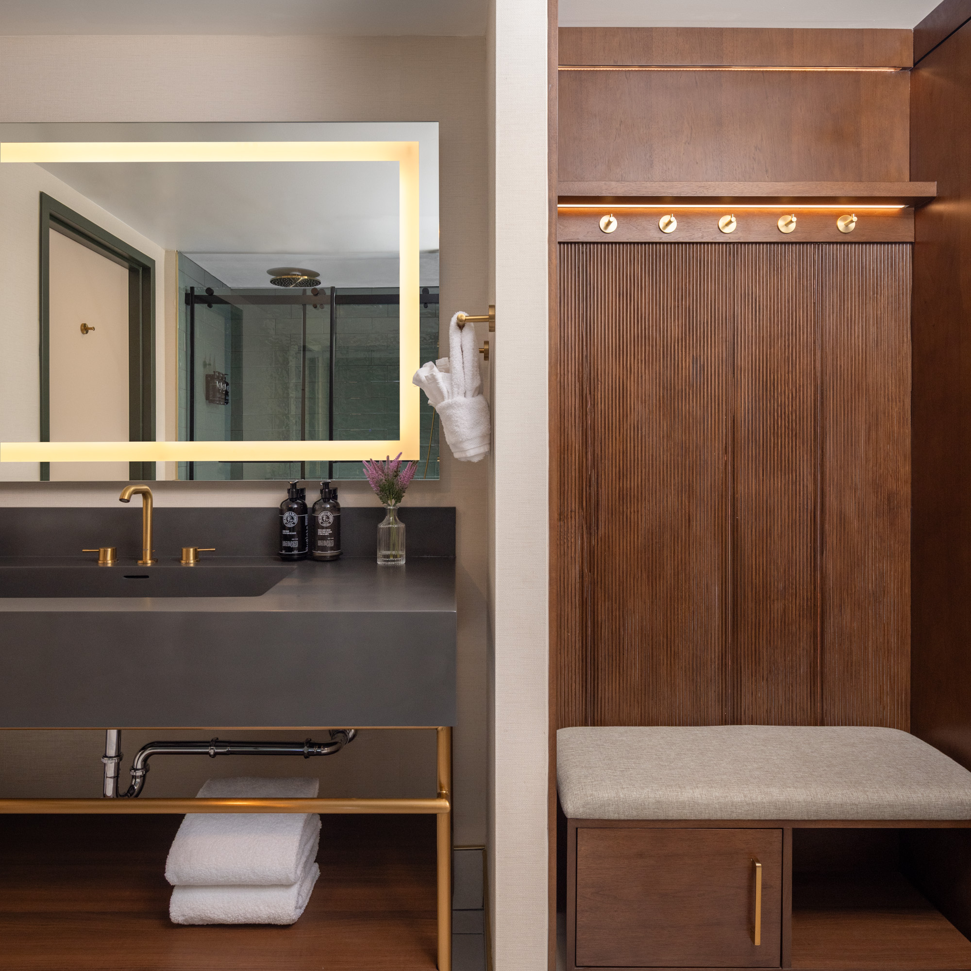 Modern bathroom with a large lighted mirror, dark countertop, gold fixtures, towels, toiletries, small flower vase, and a wooden bench with hooks.