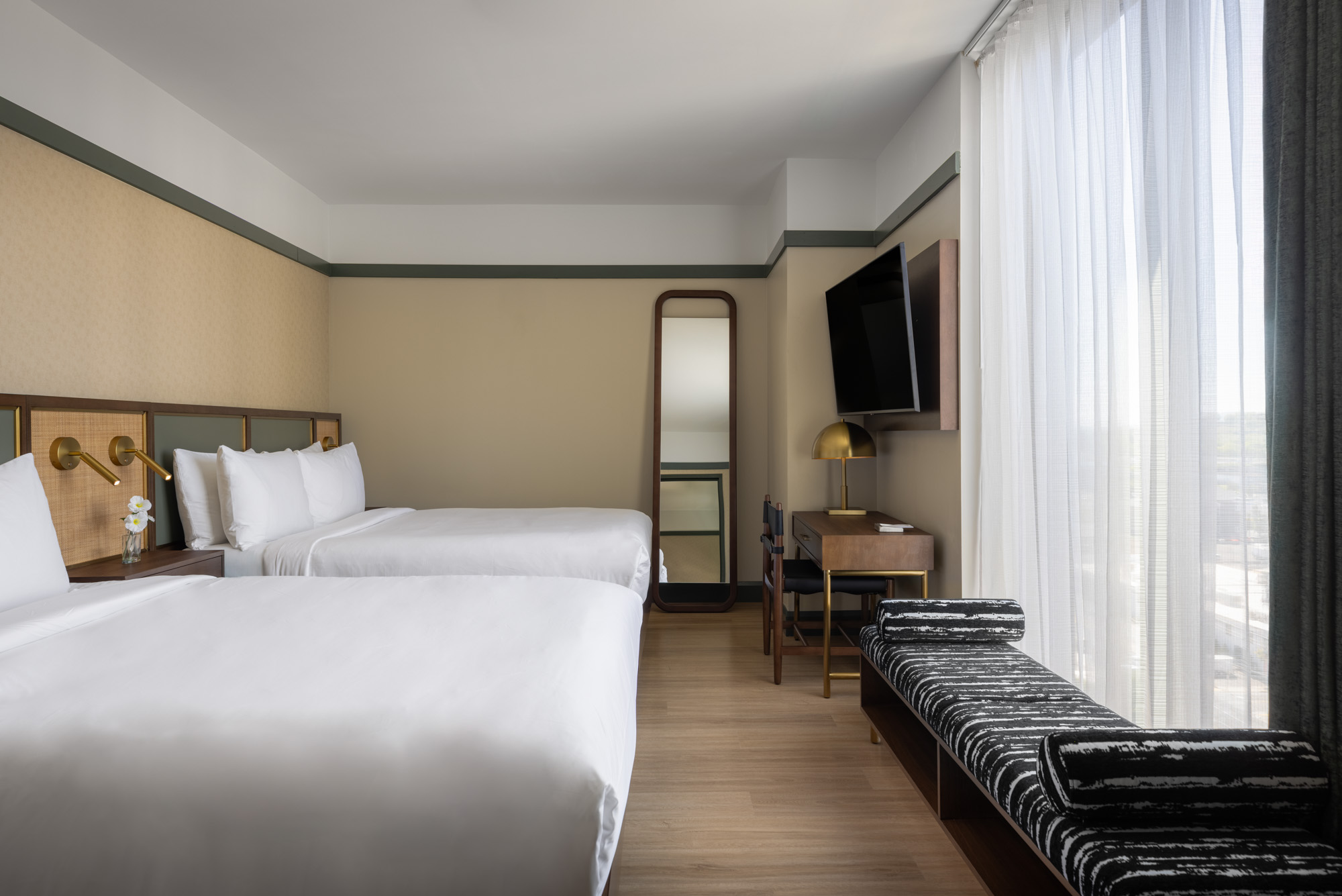 Hotel room with two double beds, a desk with a lamp and chair, a large wall mirror, wall-mounted TV, and a cushioned window bench beside floor-to-ceiling curtains.