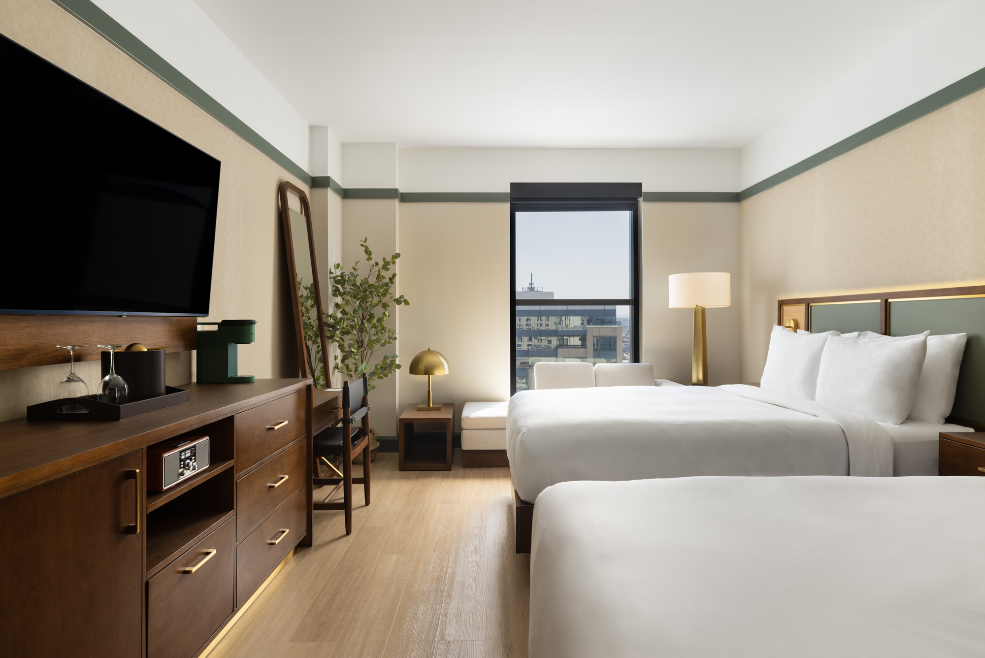 Hotel room with two double beds, a large TV, wooden furniture, a window with city view, and modern decor including a floor lamp and a potted plant.