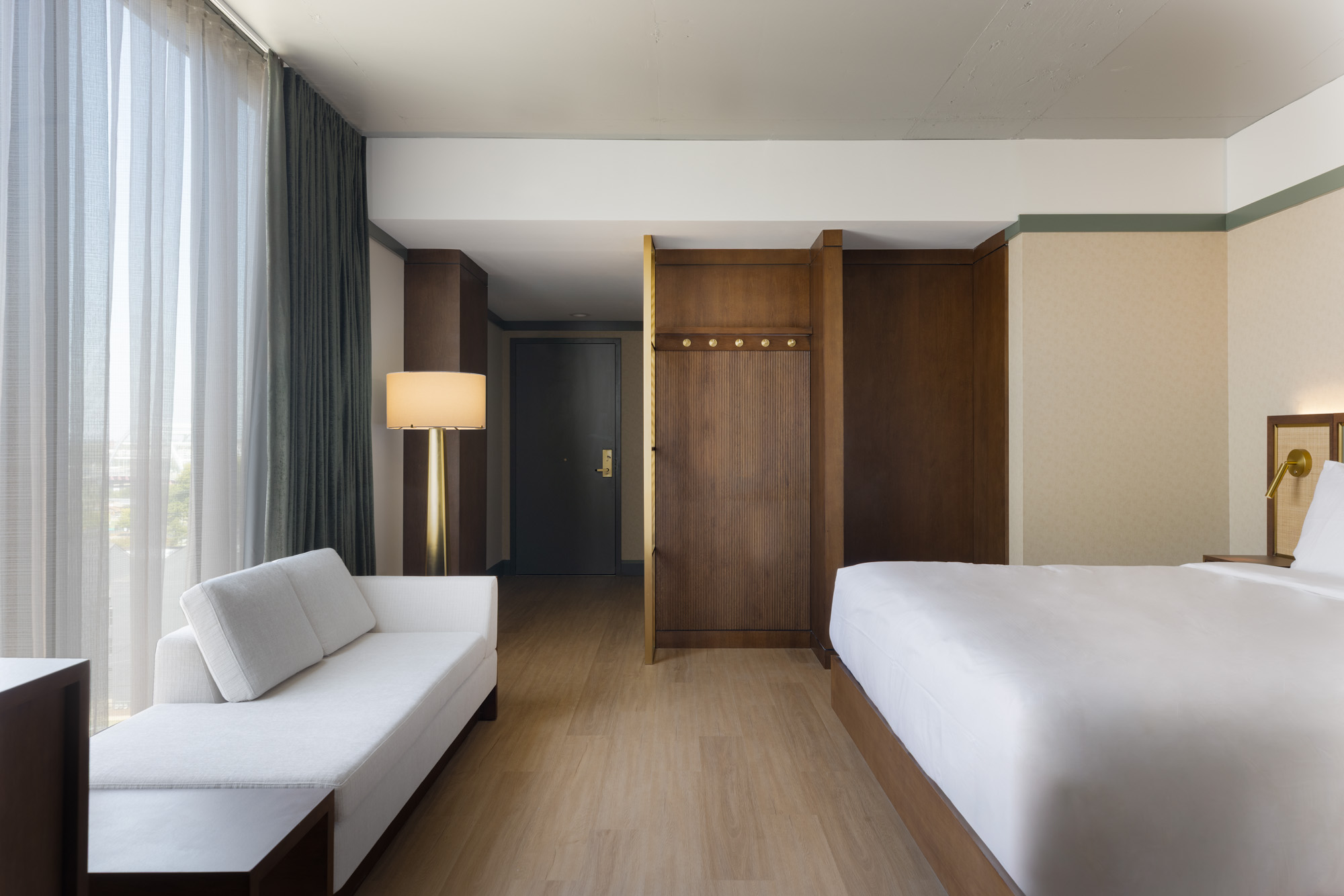 Modern hotel room with a large bed, white sofa, wooden floors, floor-to-ceiling window with curtains, lamp, and a closet near the entrance door.