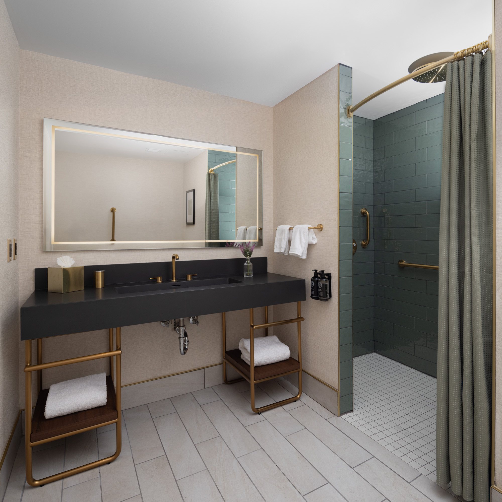 Modern hotel bathroom with a large mirror, black countertop, towels, toiletries, and a walk-in shower with a gold grab bar, green tiles, and a shower curtain.