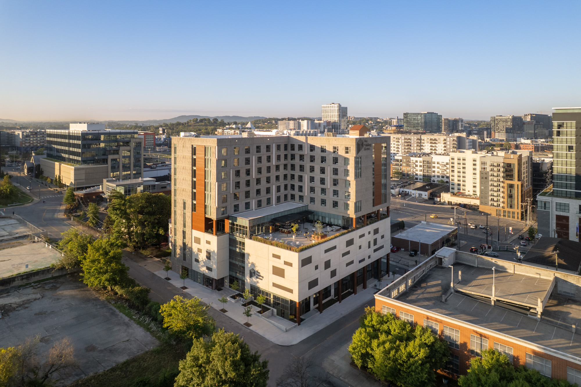 A modern mid-rise apartment building with a central courtyard and outdoor terrace, surrounded by city streets and other urban buildings under clear blue skies.