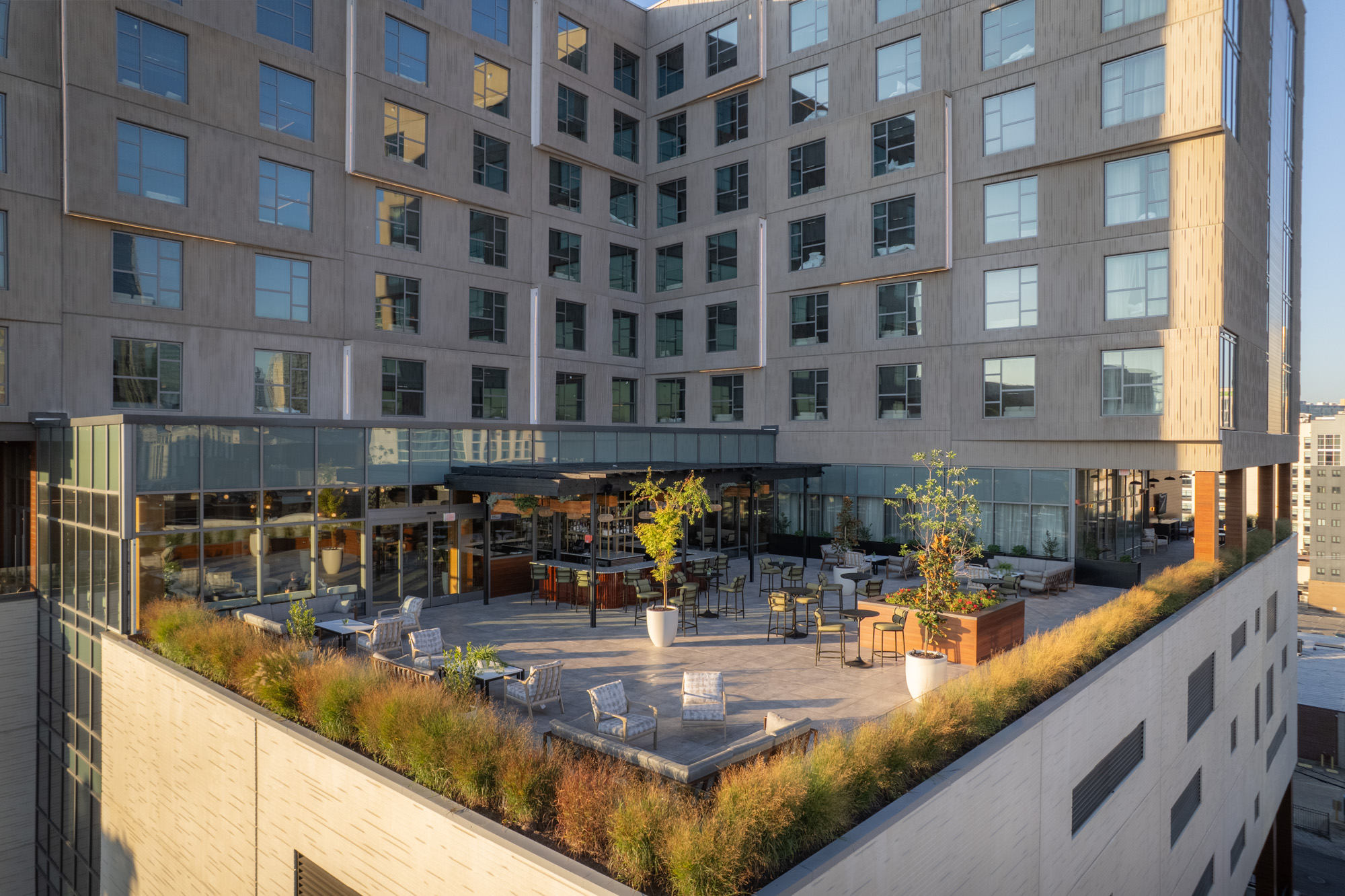 Outdoor terrace with tables, chairs, and potted plants on a modern building's rooftop, surrounded by tall windows and city views.