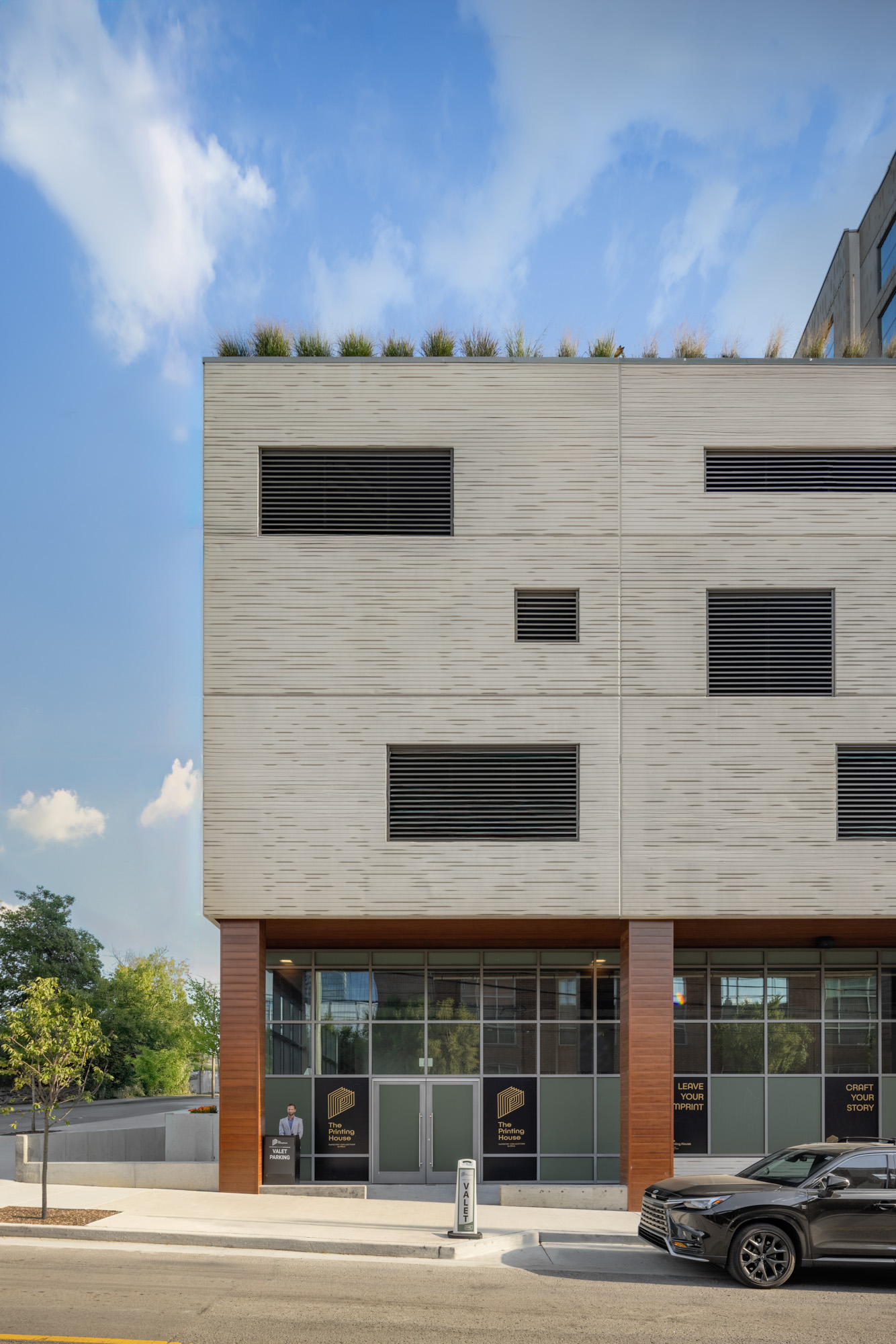 Modern three-story building with beige facade, rectangular black vents, ground-floor glass windows, and a few small trees visible nearby; car parked in front on the right.